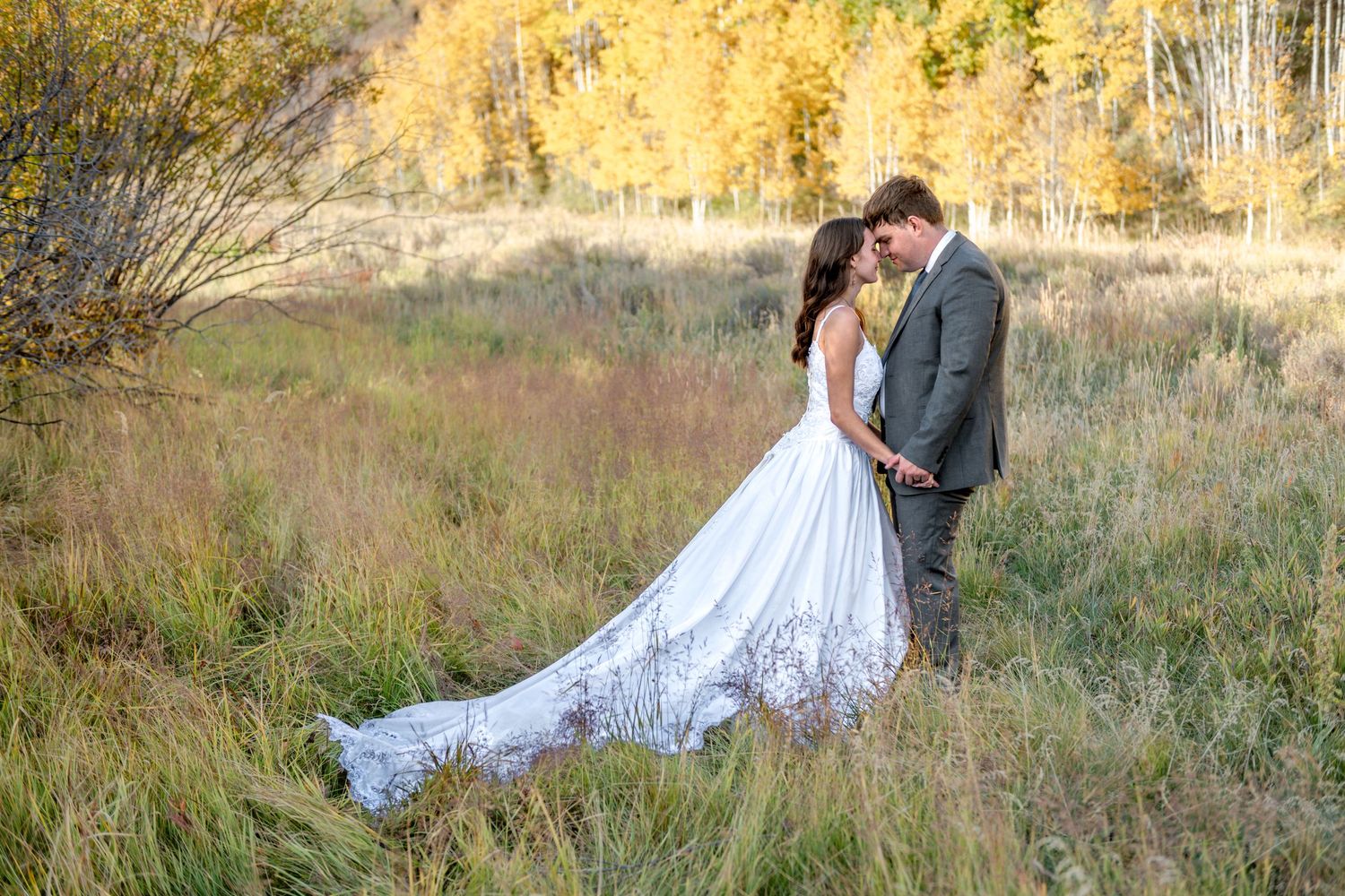 A couple in wedding attire embraces in a golden autumn field during sunset. They are facing each other and have their foreheads together.