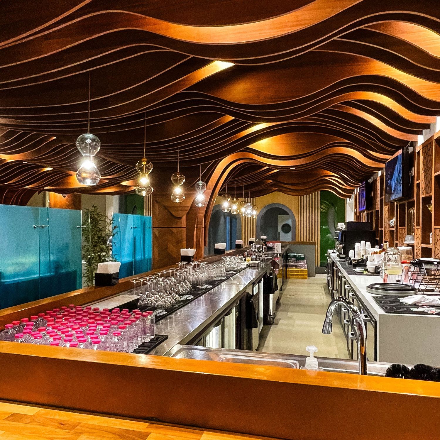 Modern restaurant bar interior with wavy wooden ceiling design, pendant lights, and sleek counter setup with glassware.