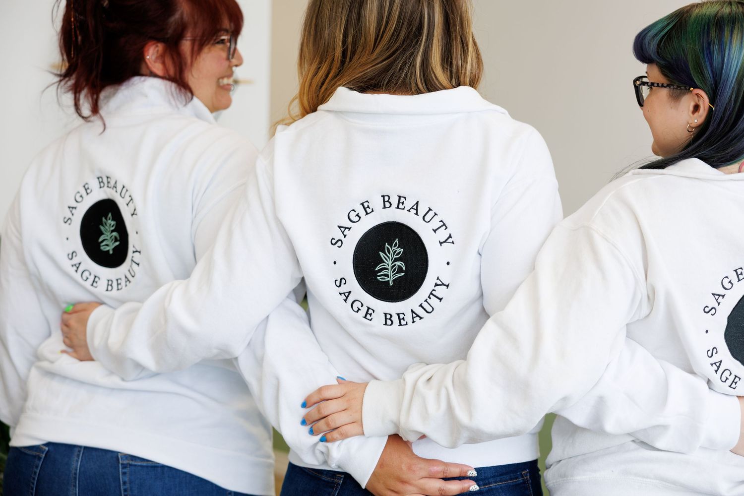 Three people wearing matching white sweatshirts with circular logo designs on the back stand together in a casual group photo.