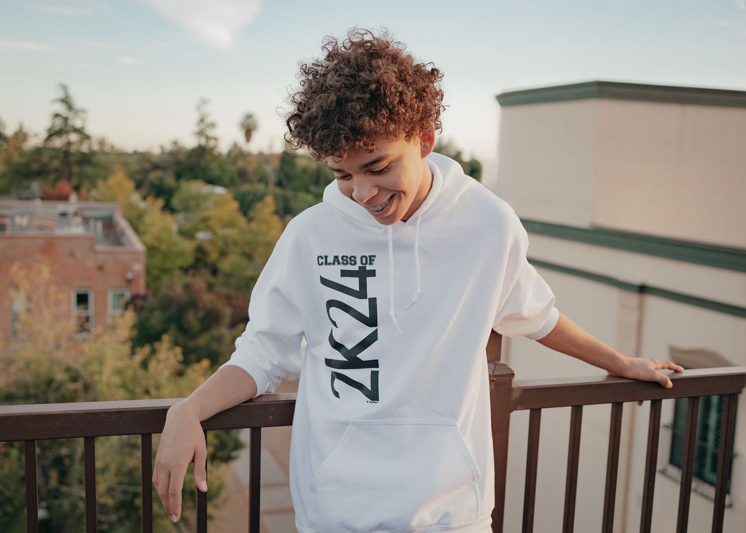 Senior boy in white Class of 2024 hoodie leaning against railing on top of a building in Winters, CA looking at the ground smiling