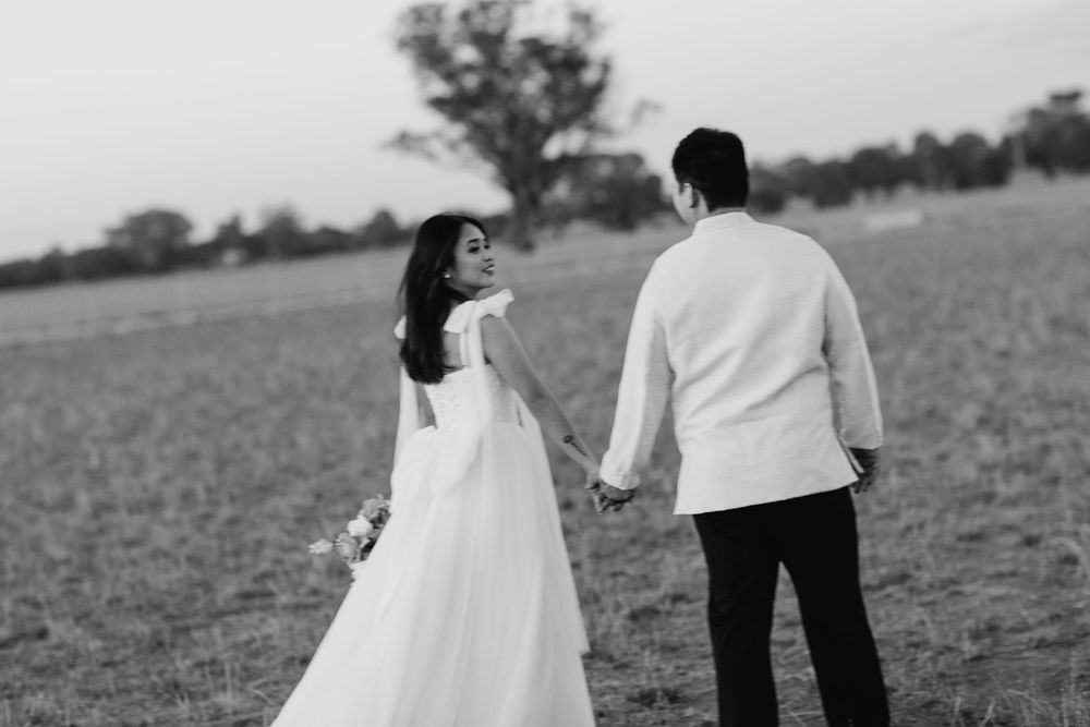 Black and white photo of couple in wedding attire holding hands while standing in an open field at sunset.