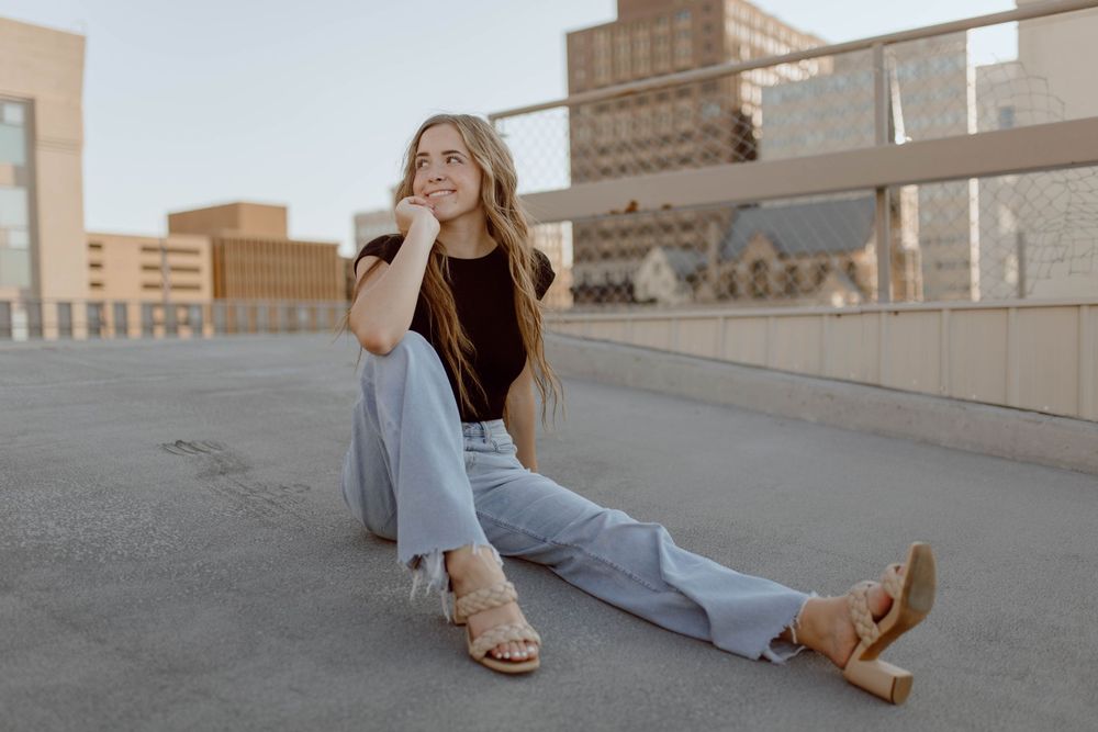 A person wearing a black tank top and light jeans sits on a rooftop at sunset with a city backdrop.