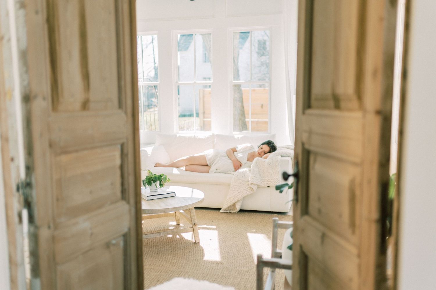 View through elegant white french doors into bright sunlit bedroom with someone resting in crisp white bedding.