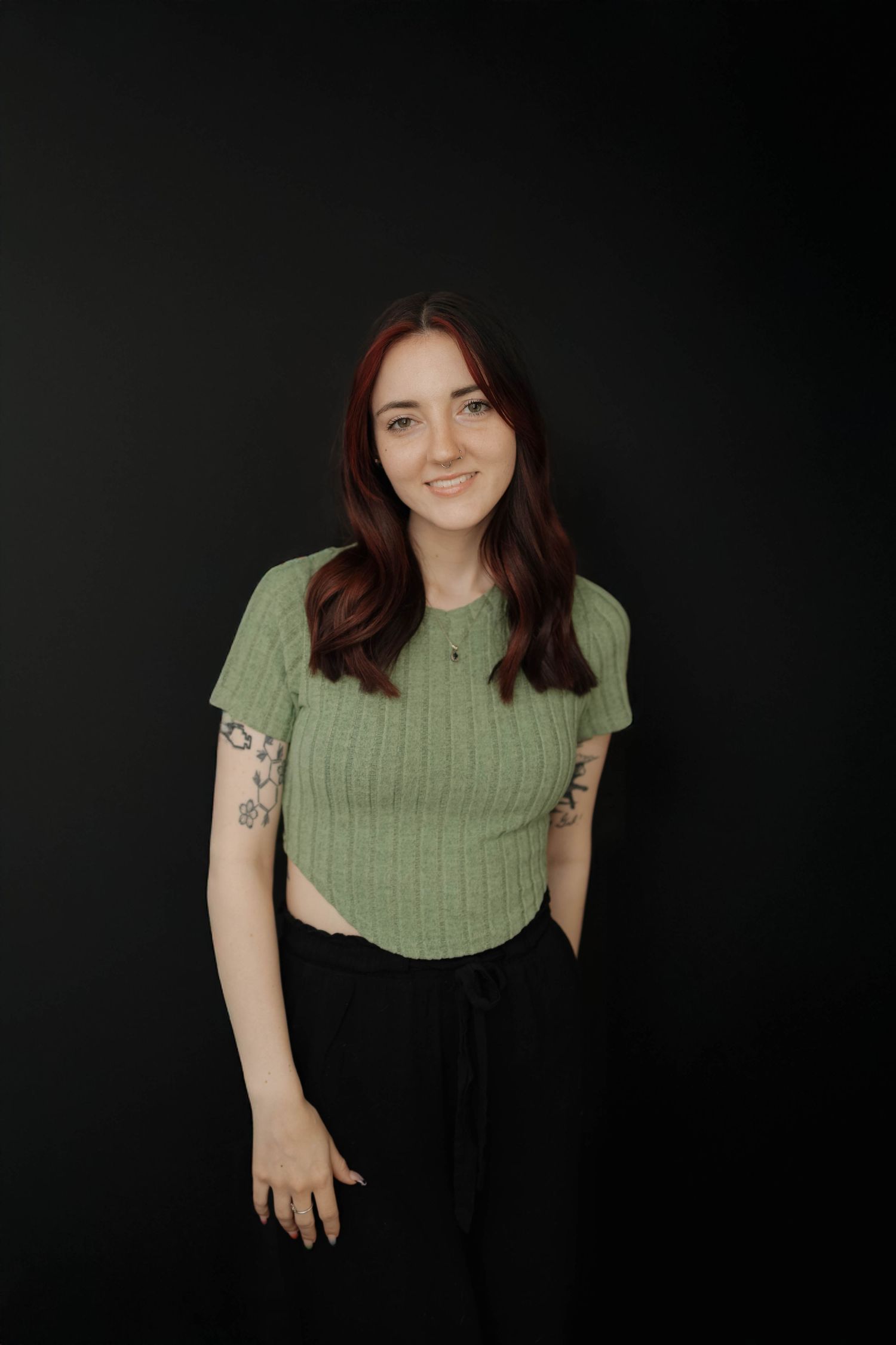 Studio portrait against black background wearing a sage green ribbed top and black pants with visible arm tattoos.