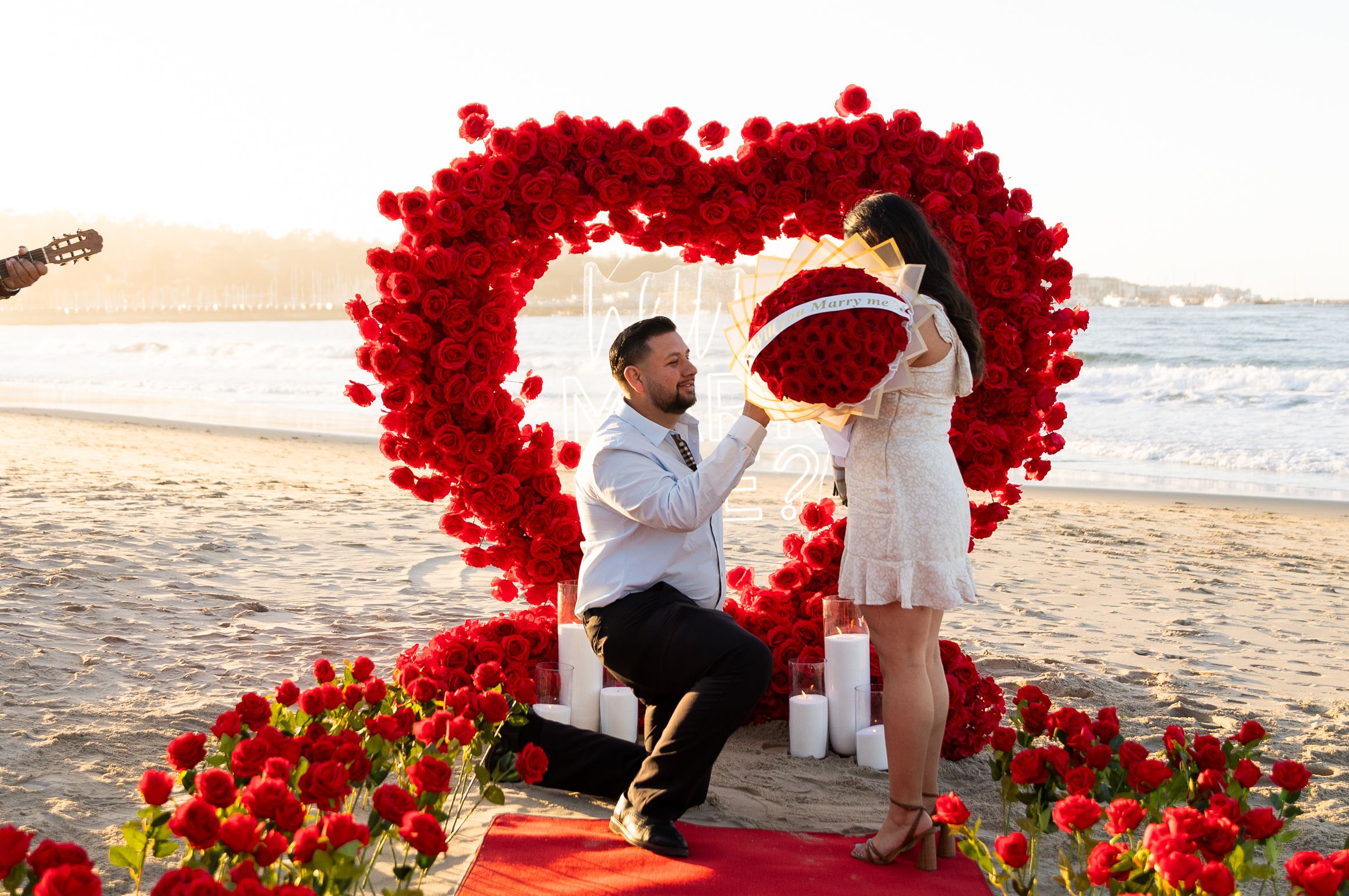 A romantic beach proposal setup with a large heart made of red roses on the sand during sunset.