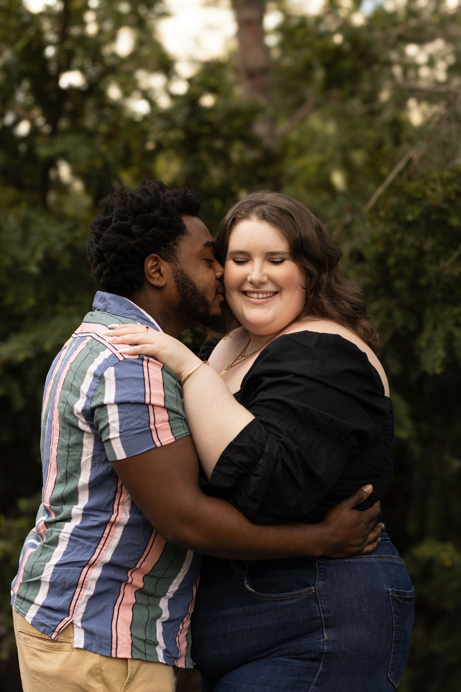 A man with a striped shirt embraces and kisses a smiling woman with long brown hair and a black top on the cheek.