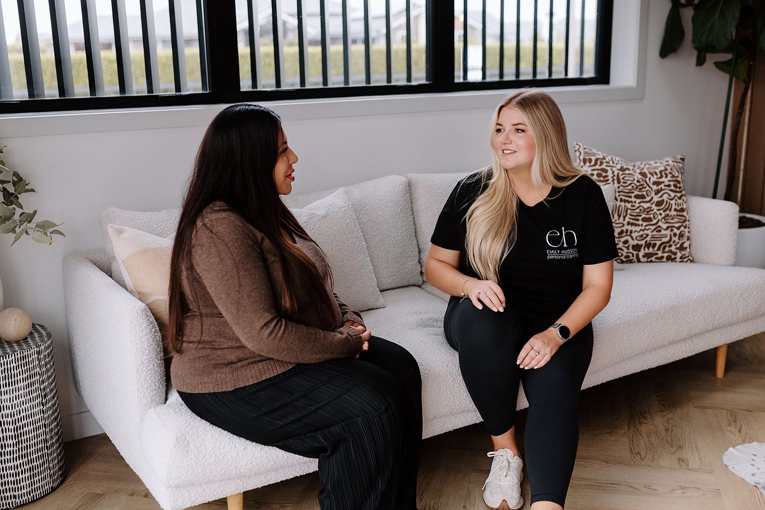 Two women sitting on a white couch having an animated conversation in a bright modern living room with decor accents.