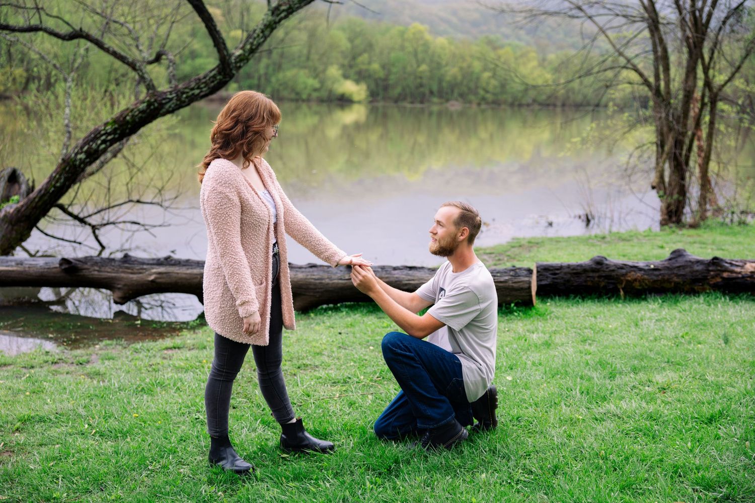 A romantic proposal scene takes place on green grass near a serene lake with trees in the background. Cove Lake Park.