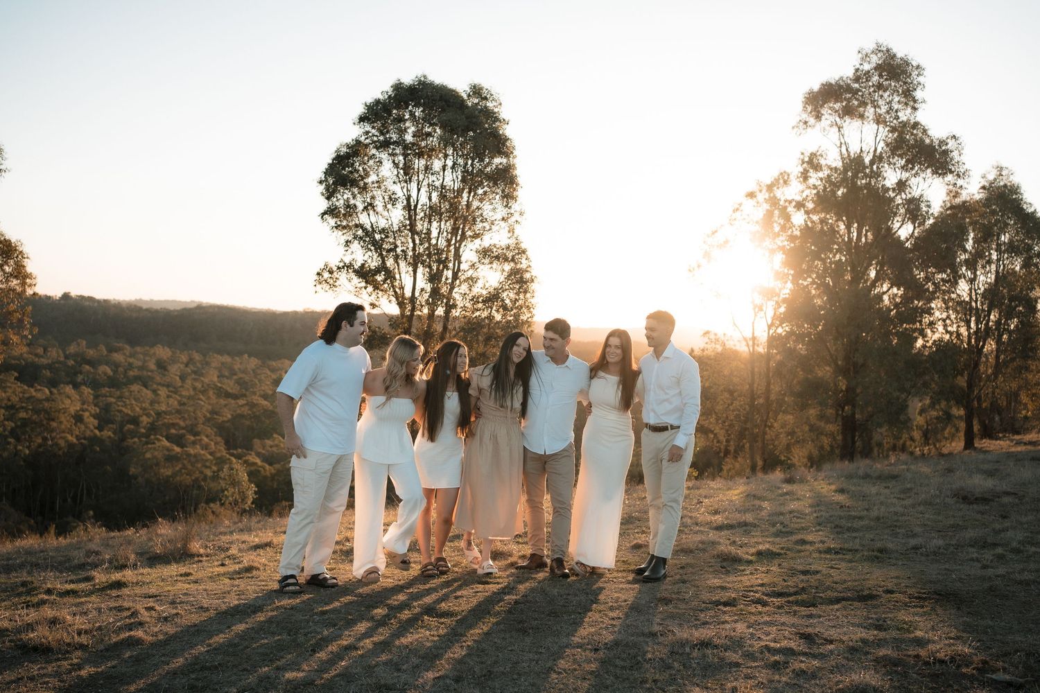 Group of people in white outfits standing together on a hilltop at golden sunset with eucalyptus trees in background.