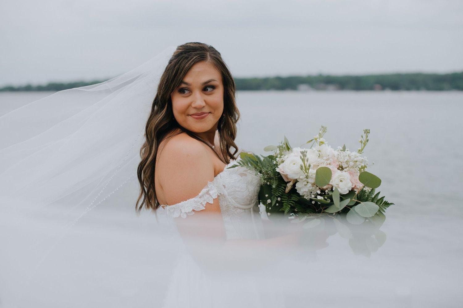 A romantic bridal portrait features an off-shoulder white dress and elegant white floral bouquet against a misty backdrop.