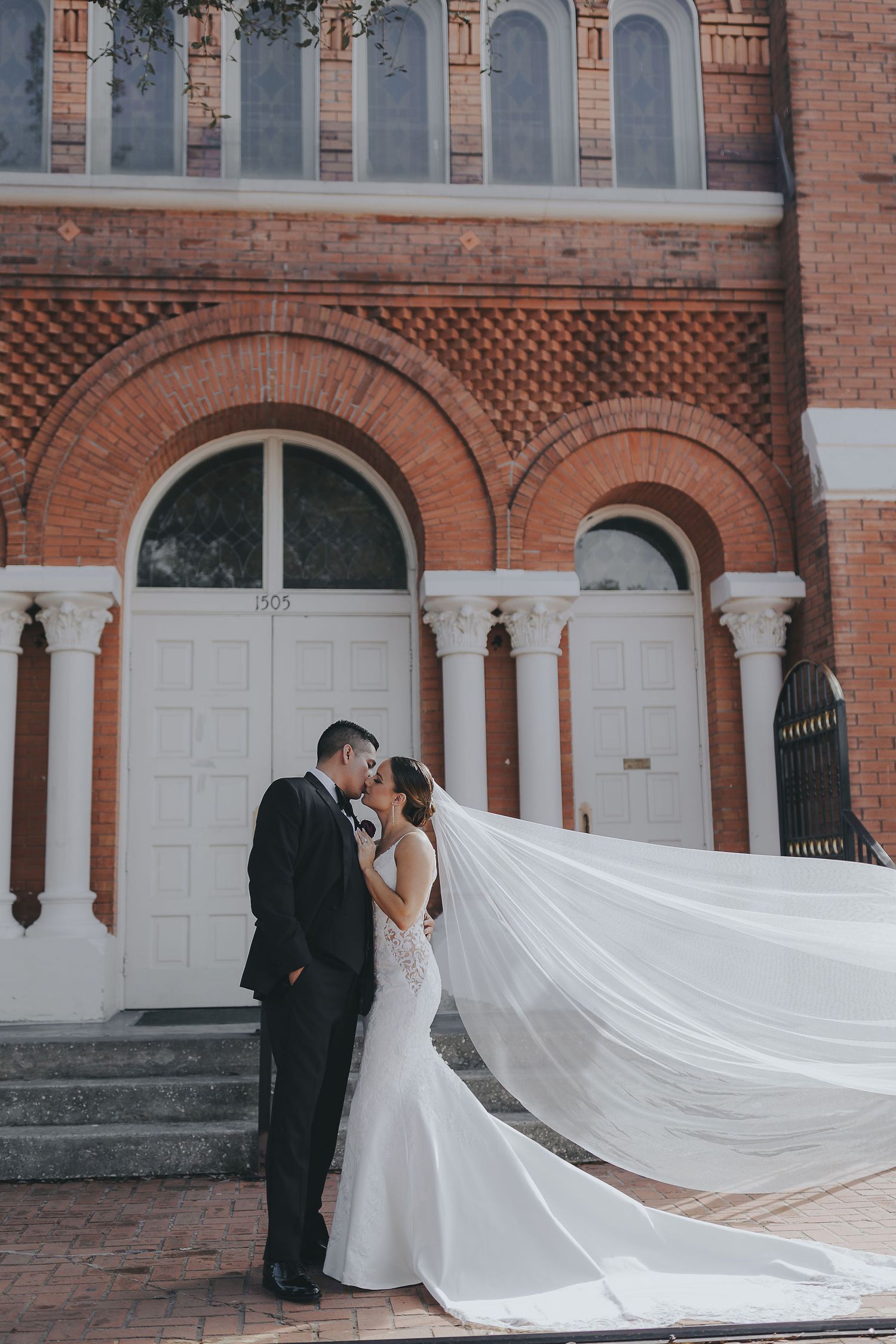 A couple embraces in front of an ornate brick church with white columns and arched doors.