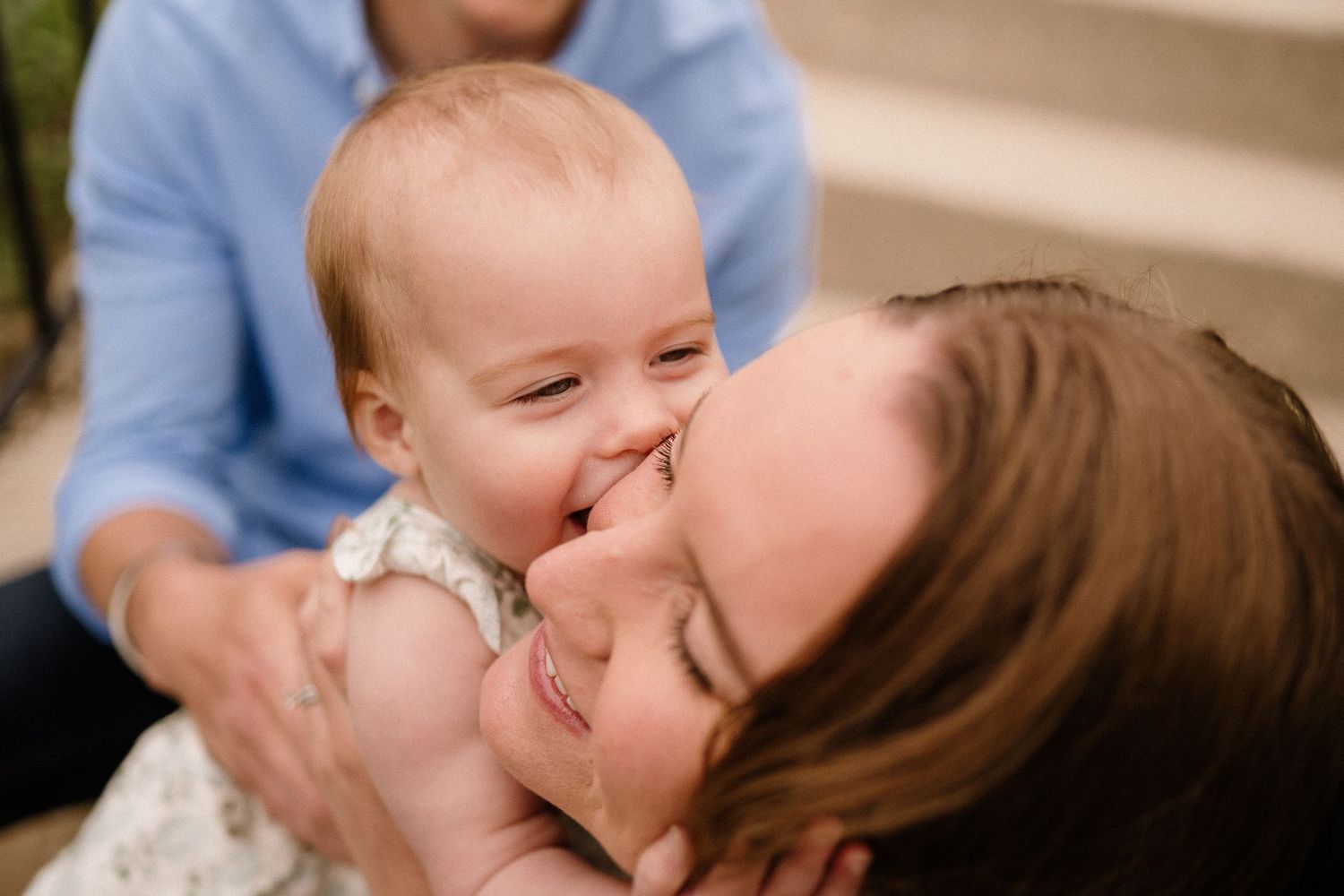 A joyful baby in a white dress smiles and cuddles close with loved one during a tender family moment.