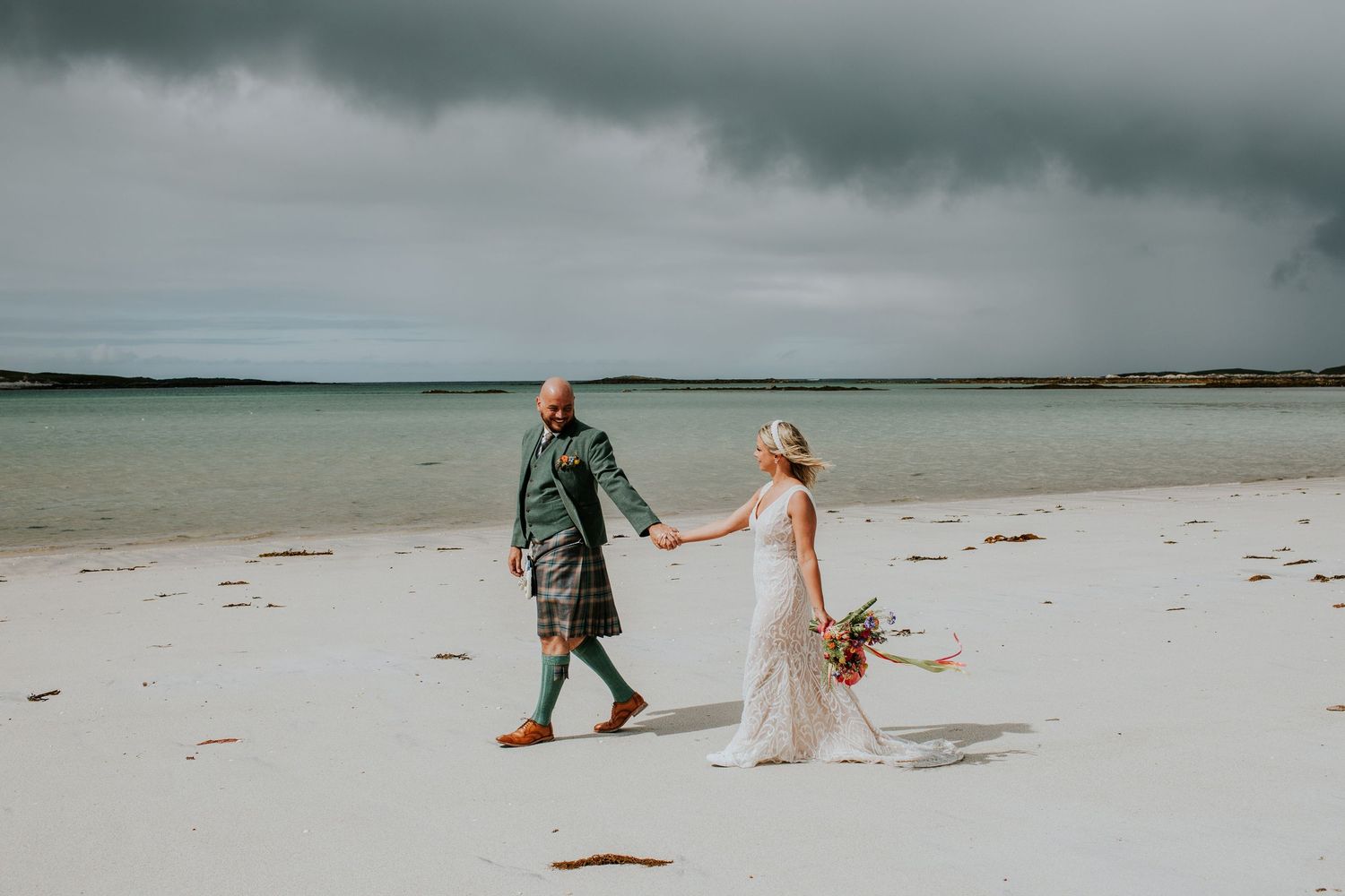 A couple walks hand in hand on a white sandy beach under stormy gray skies with a dried flower bouquet.