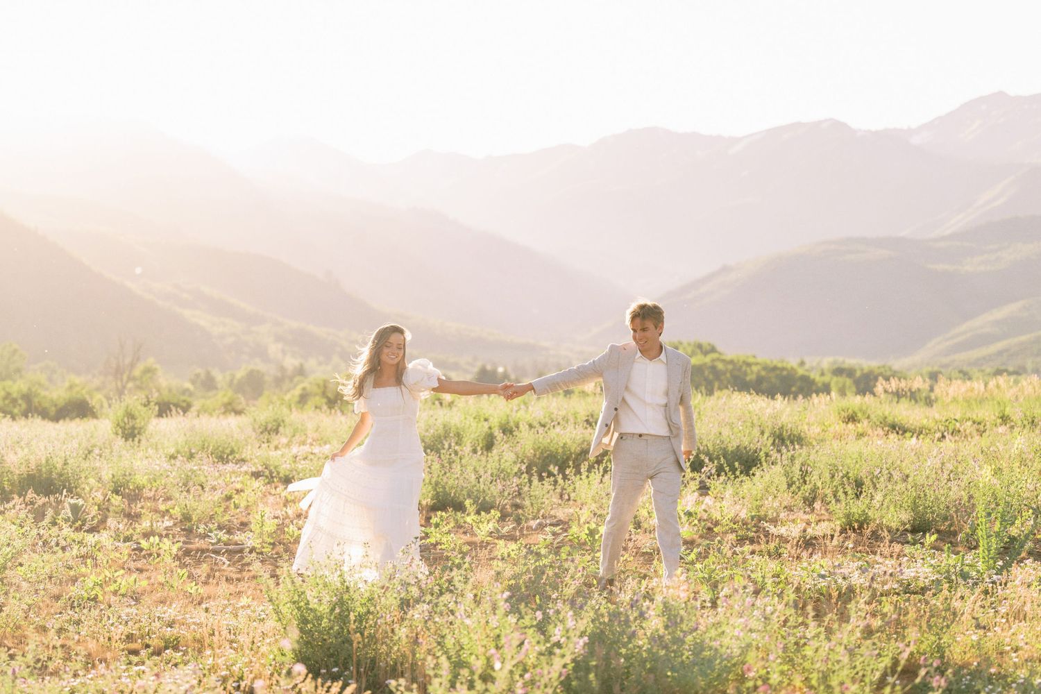 A couple holds hands in a sunlit meadow with mountains in the background during a romantic sunset photoshoot.