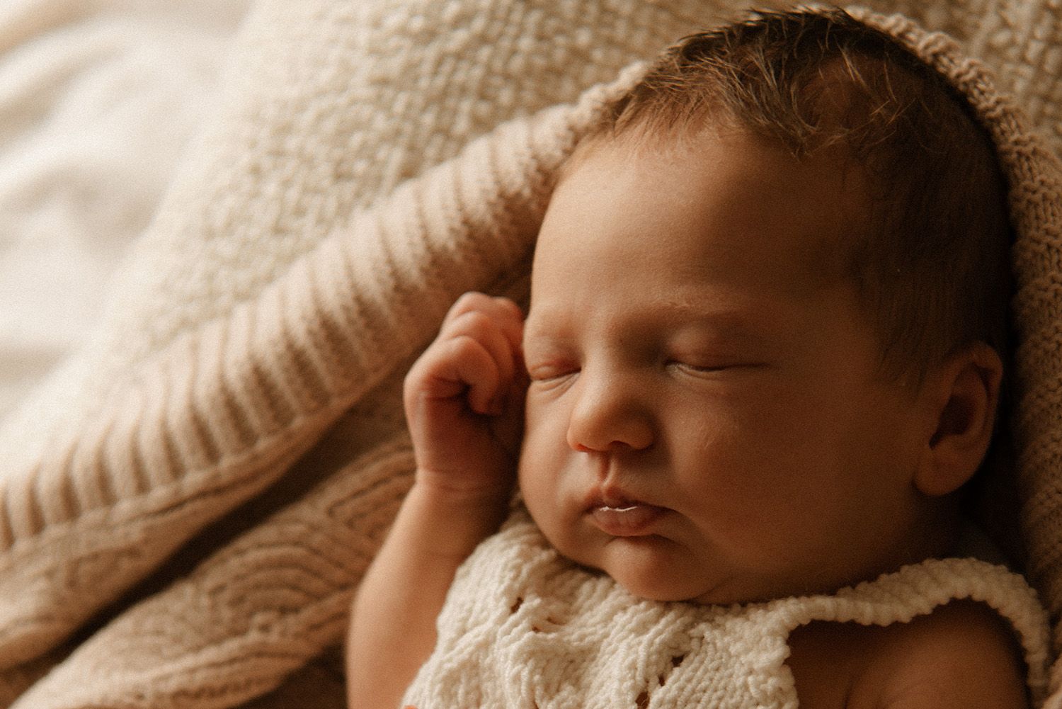 A sleeping newborn baby lies peacefully in a cozy cream-colored blanket while being held tenderly.