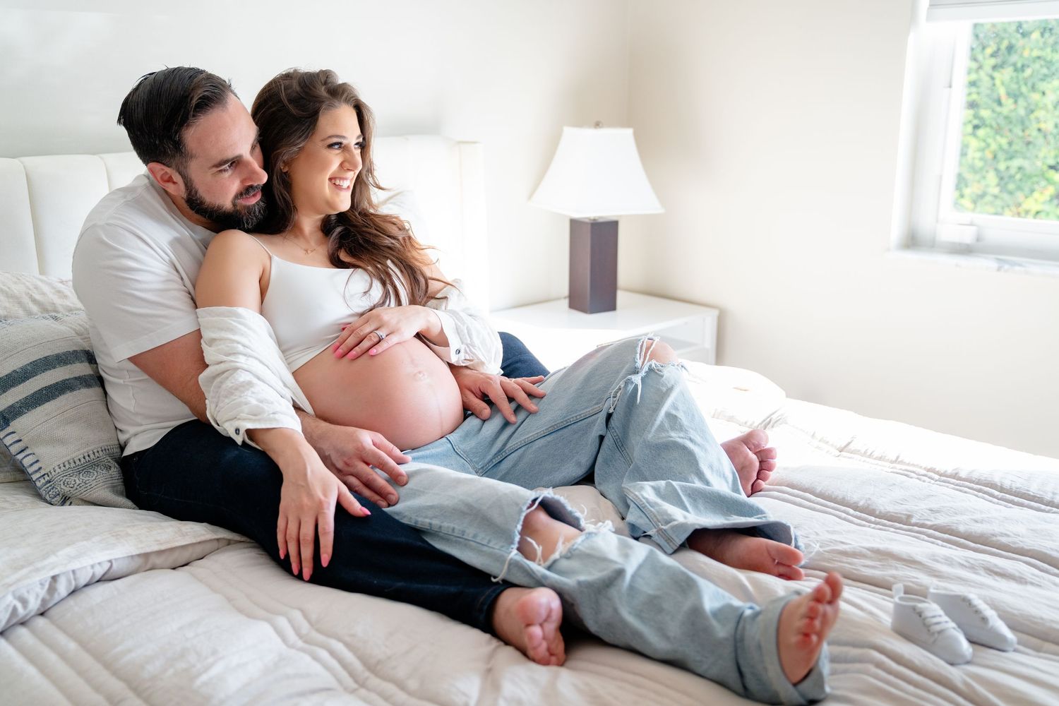 A joyful couple relaxing on bed together while tenderly cradling a pregnant belly in a sunlit bedroom.