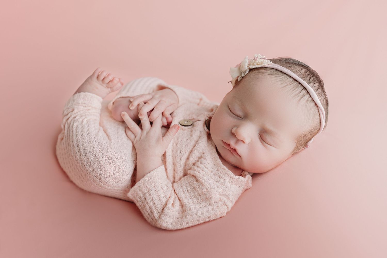 Sleeping newborn baby wearing soft pink knitted outfit and delicate headband on pink background.