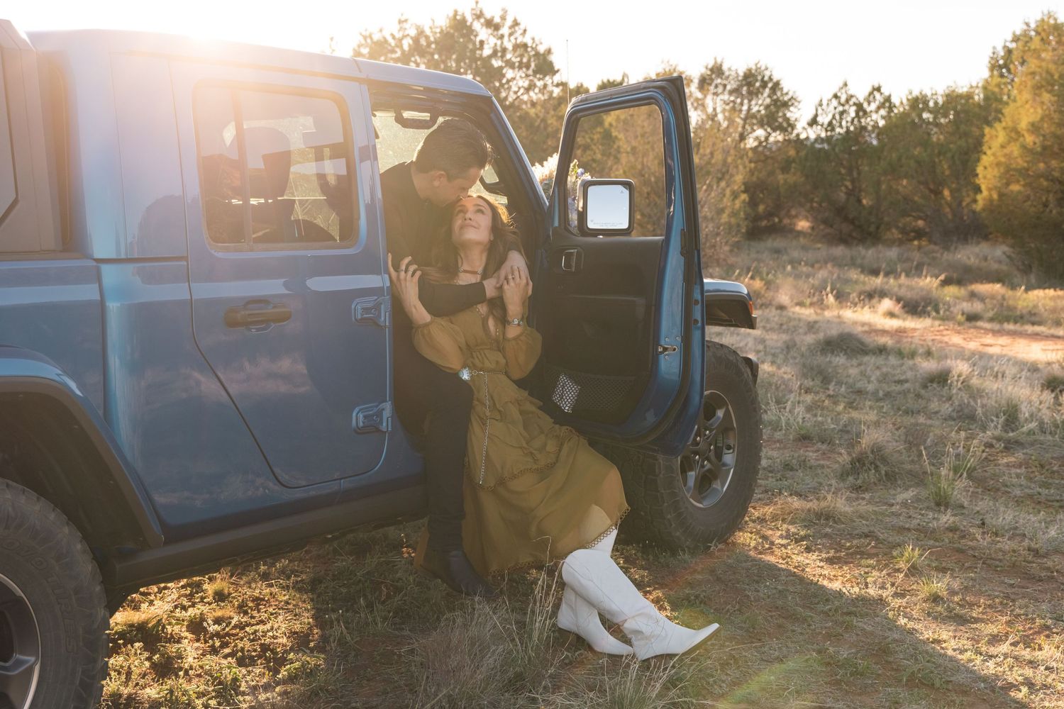 Someone in a brown jacket and white boots sits in the doorway of a blue off-road vehicle at sunset in a desert landscape.
