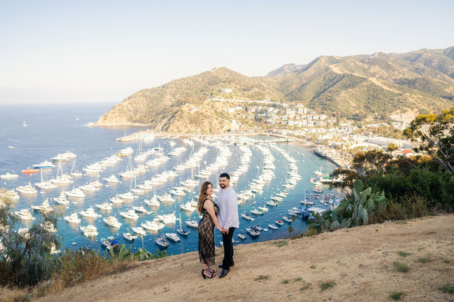 A couple stands on a cliff overlooking a marina filled with boats in Catalina Island.