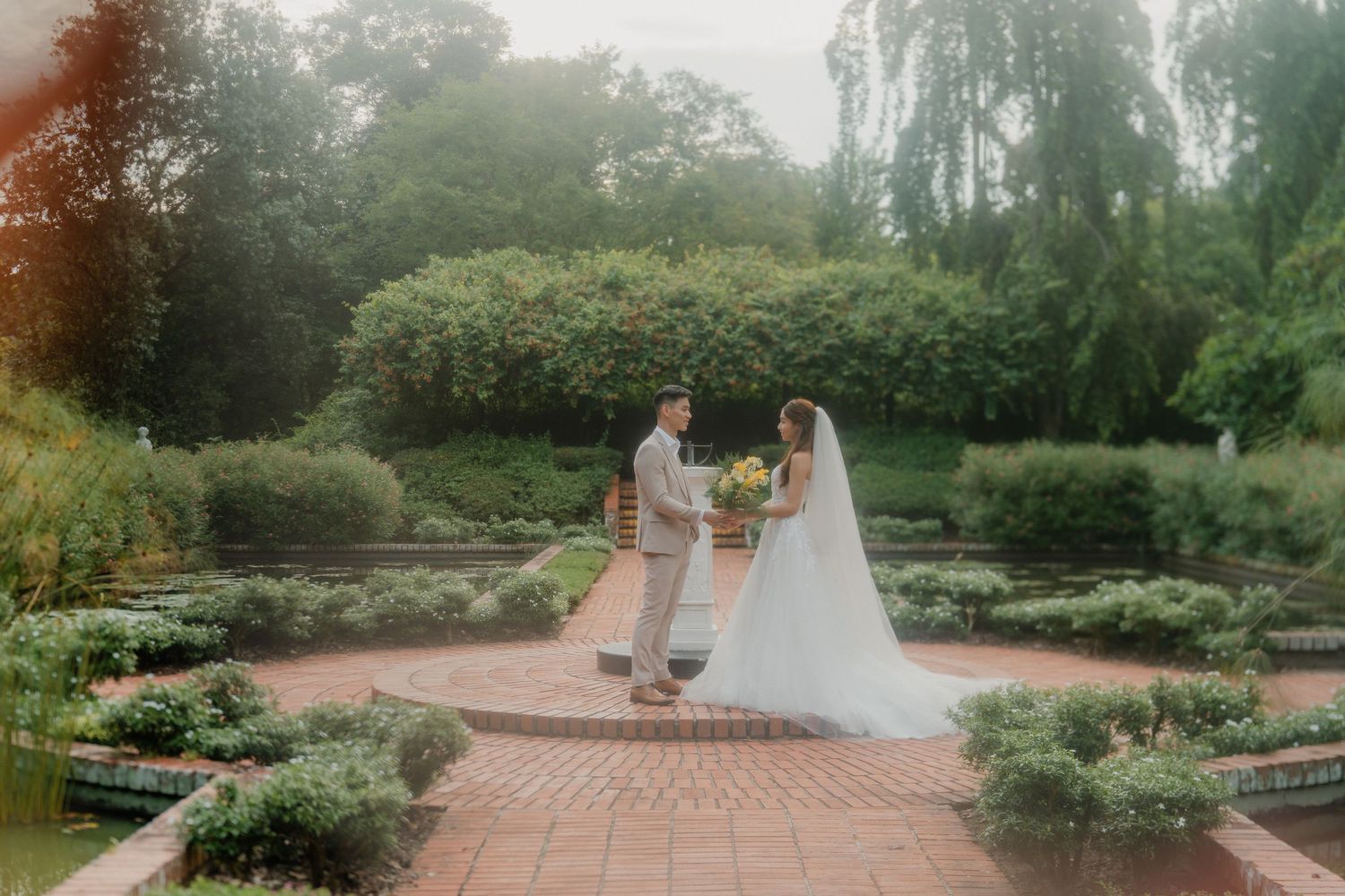 A couple embraces on a garden path beside a tranquil pond surrounded by lush greenery and blooming flowers.