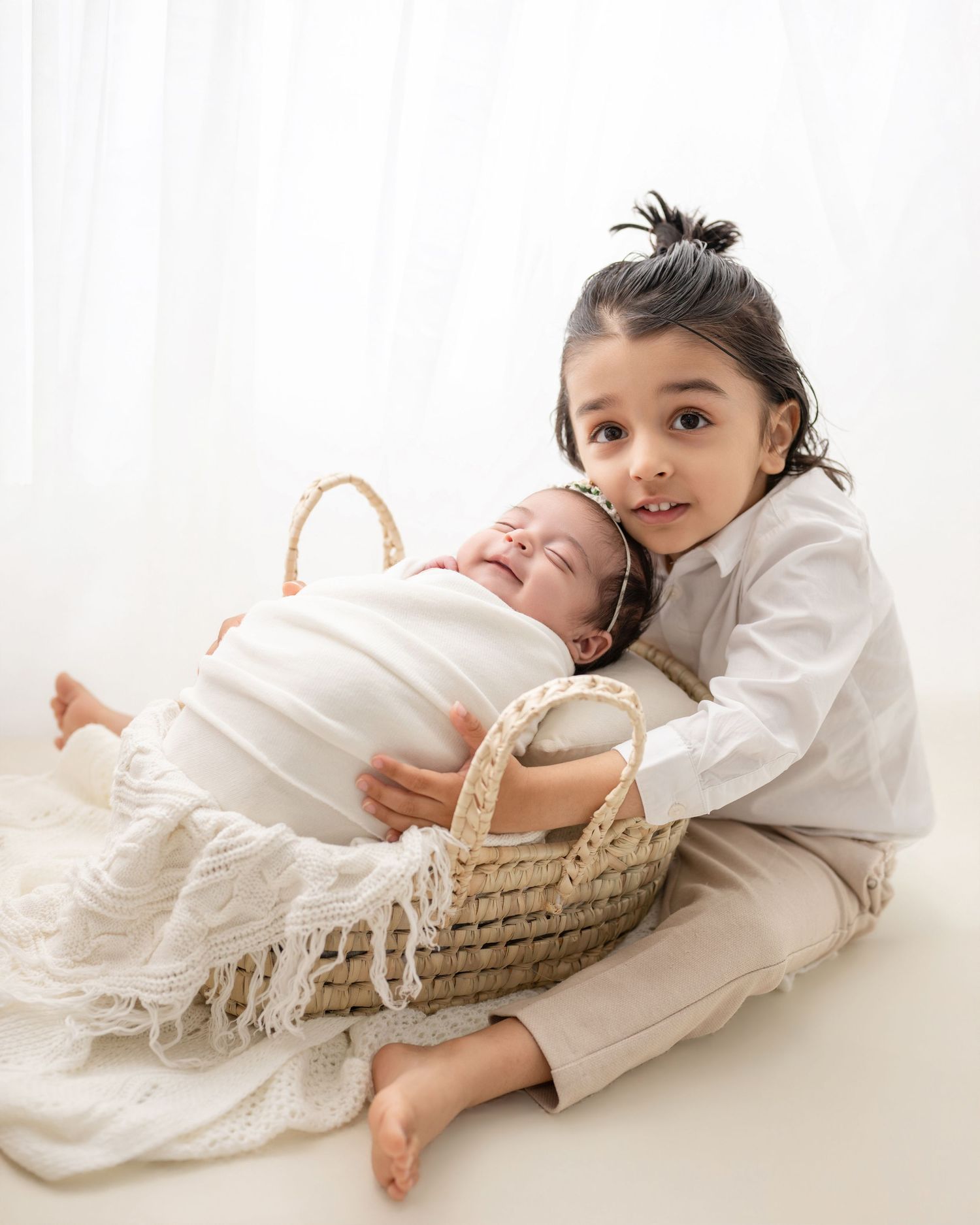 Older child in white shirt hugs swaddled newborn baby in a wicker basket during a sibling newborn photo session.