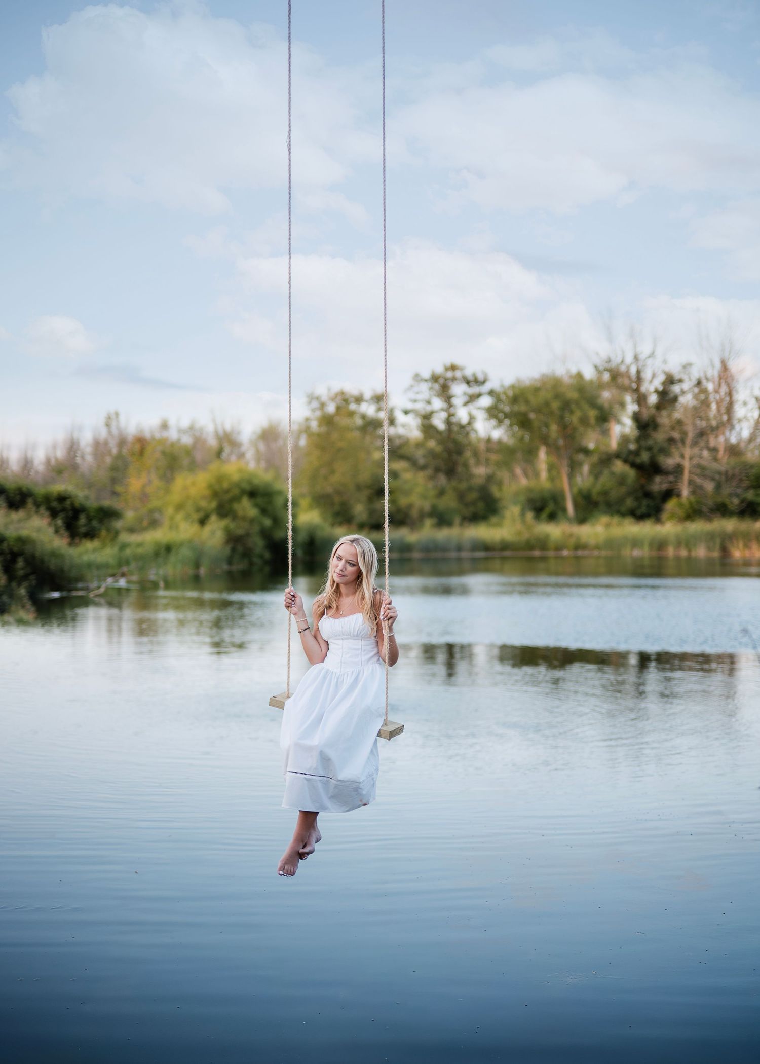 A senior girl in a white dress sits on a swing over a pond for senior photos on her family property in Sheboygan County, WI