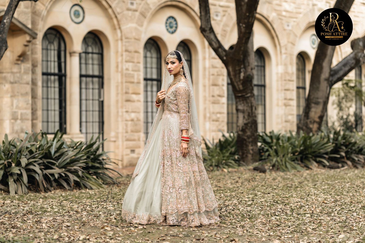 Bride in elegant embroidered lehenga poses gracefully outdoors near stone building, wearing traditional South Asian bridal attire.