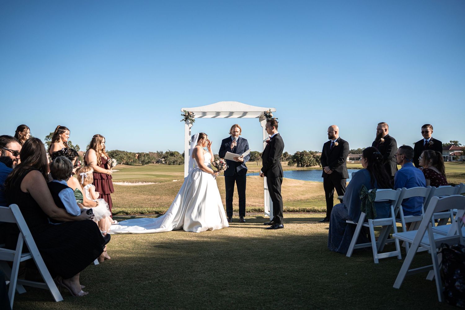 Outdoor wedding ceremony on a golf course with bride and groom at floral arch, wedding party, and seated guests under clear blue sky.