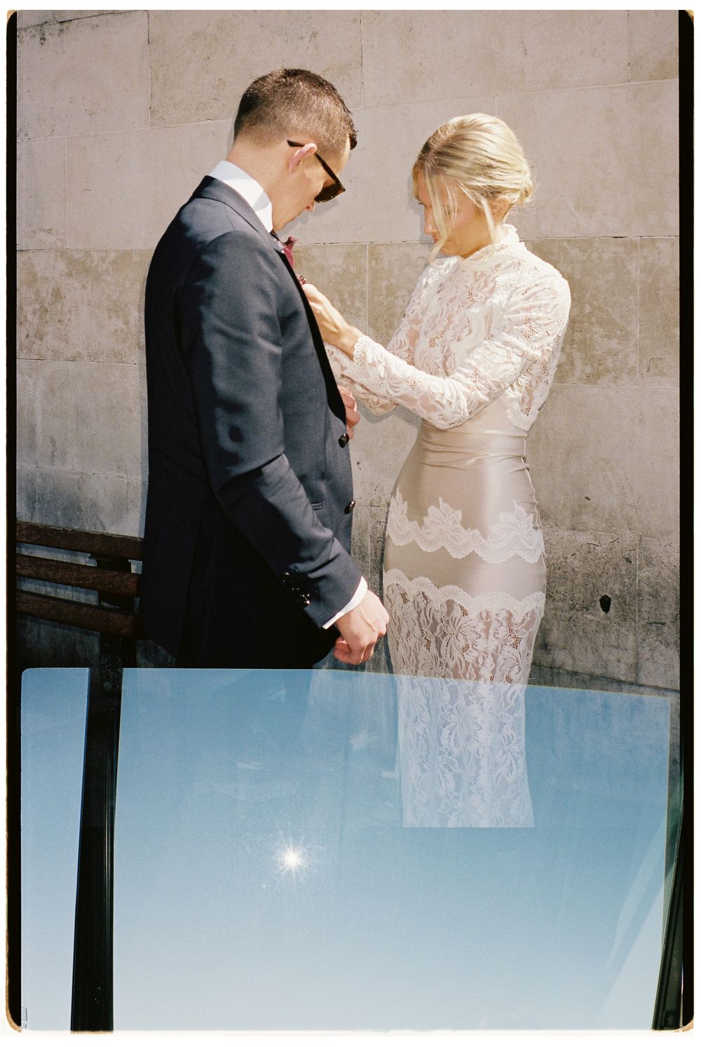 Bride in lace dress adjusting grooms suit jacket outdoors on their wedding day.