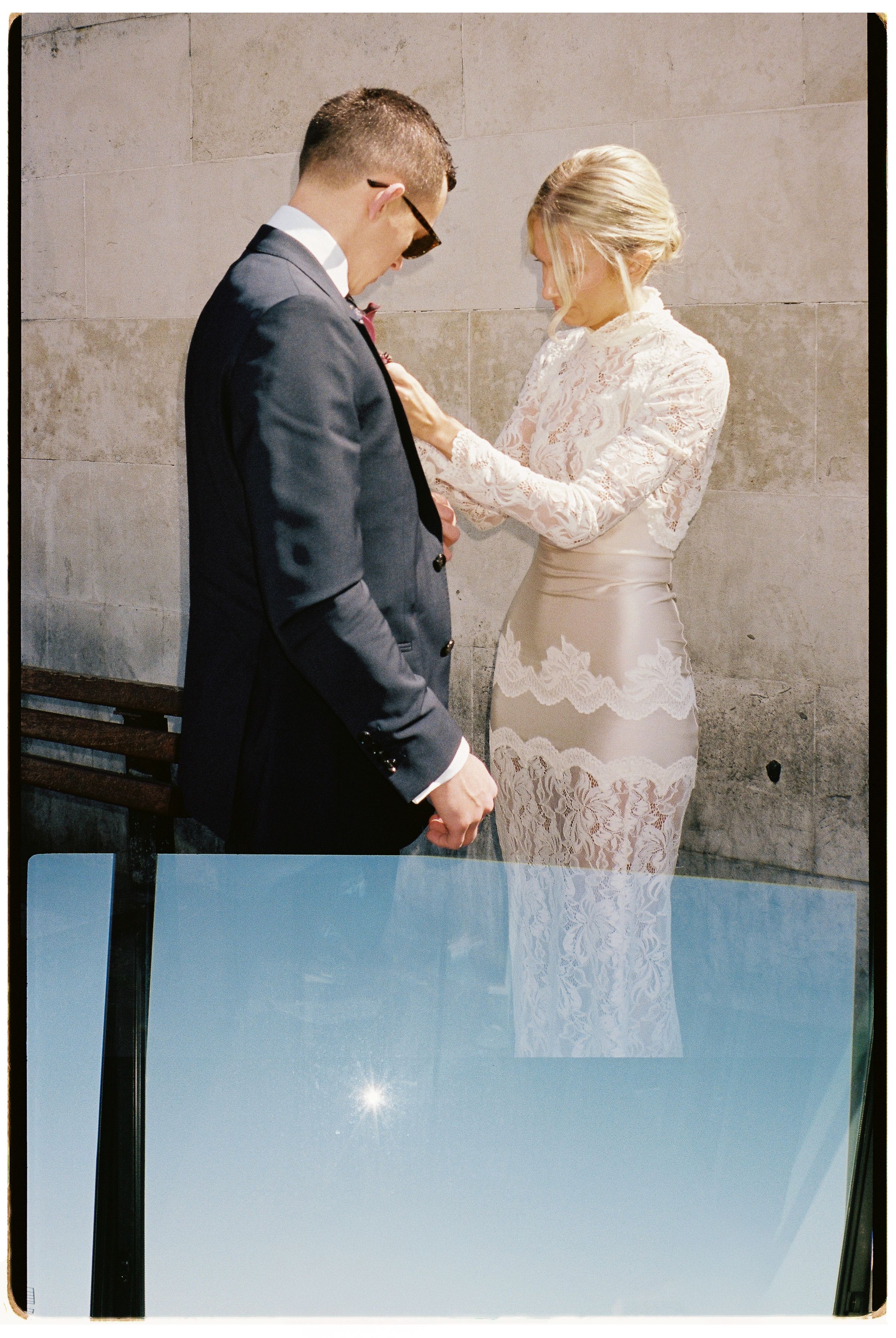 Bride in lace dress adjusting grooms suit jacket outdoors on their wedding day.