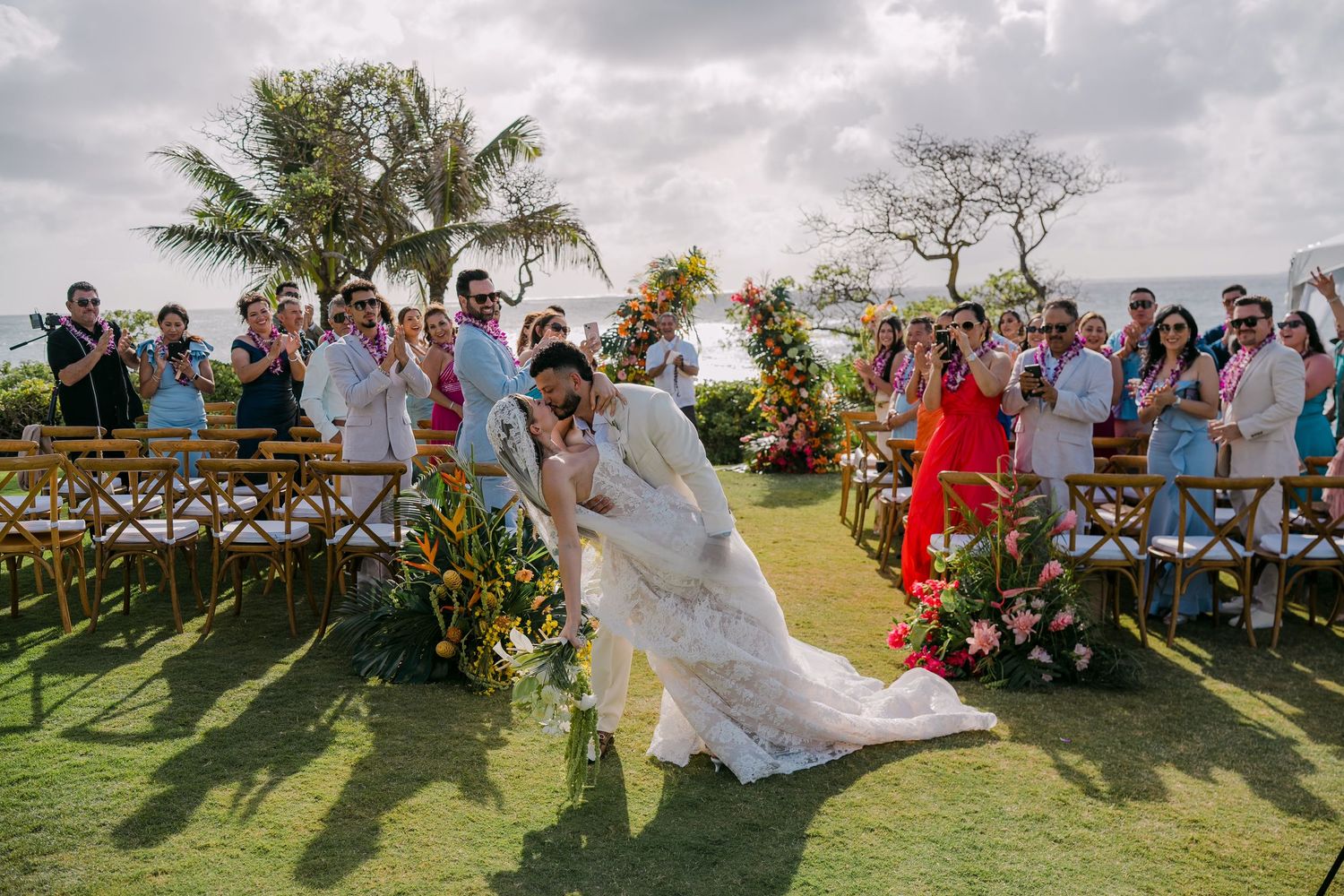 Bride and groom share a romantic dip kiss at an outdoor wedding ceremony with guests cheering on a tropical lawn.