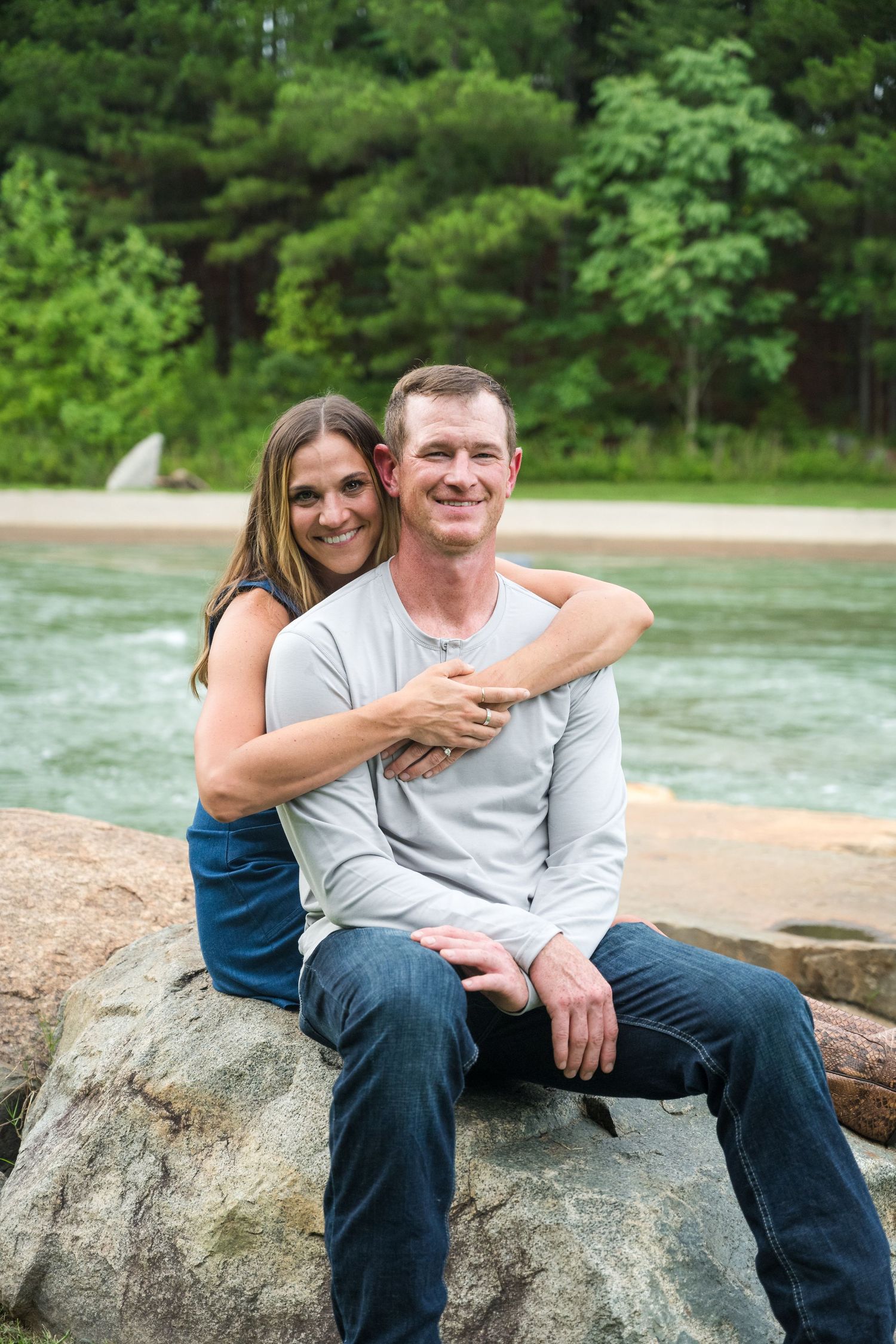 A smiling couple poses on rocks by a river, woman hugging man from behind, with lush green trees in background.