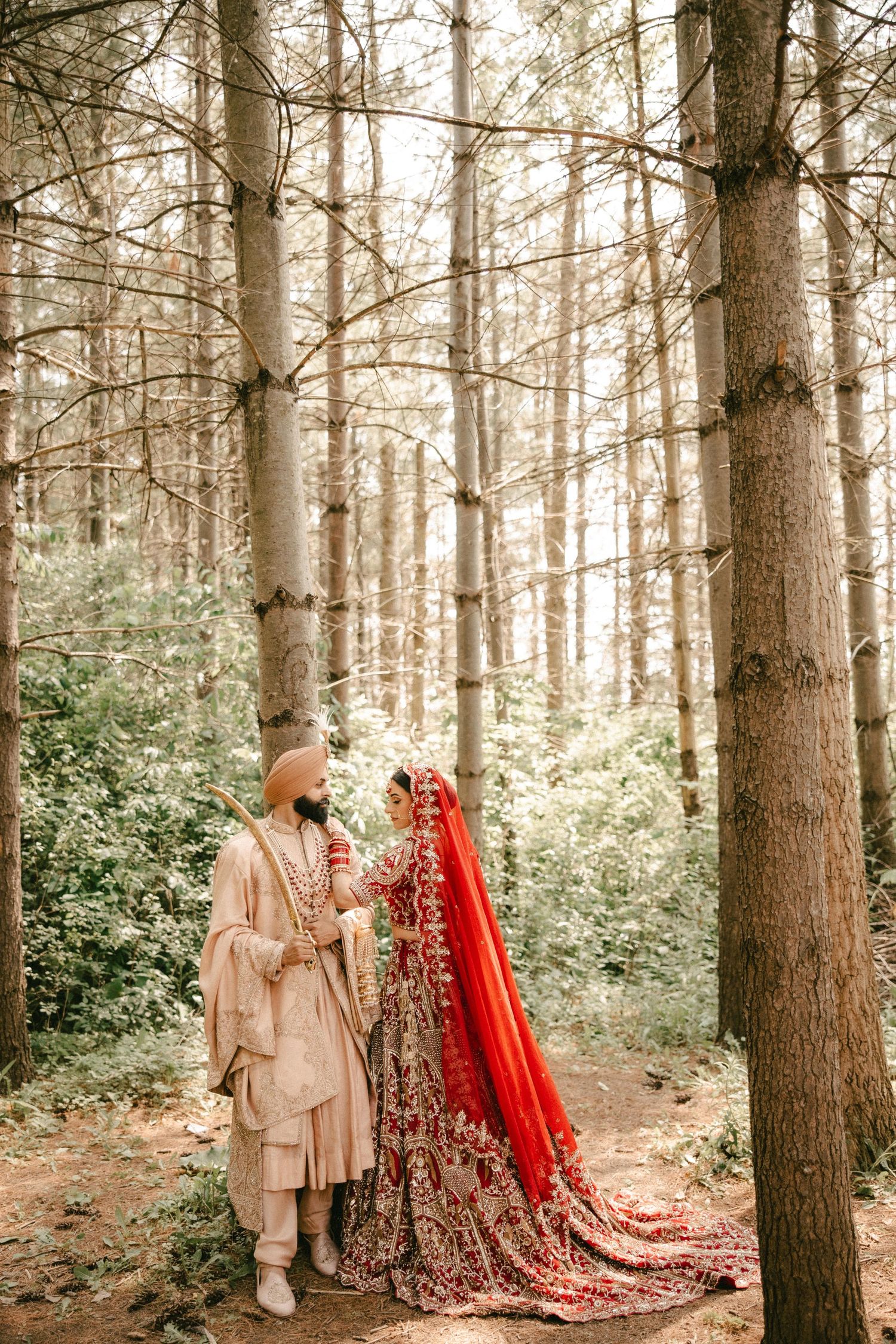 Sikh bride in red lehenga and groom in beige sherwani share a romantic moment in a serene forest setting.
