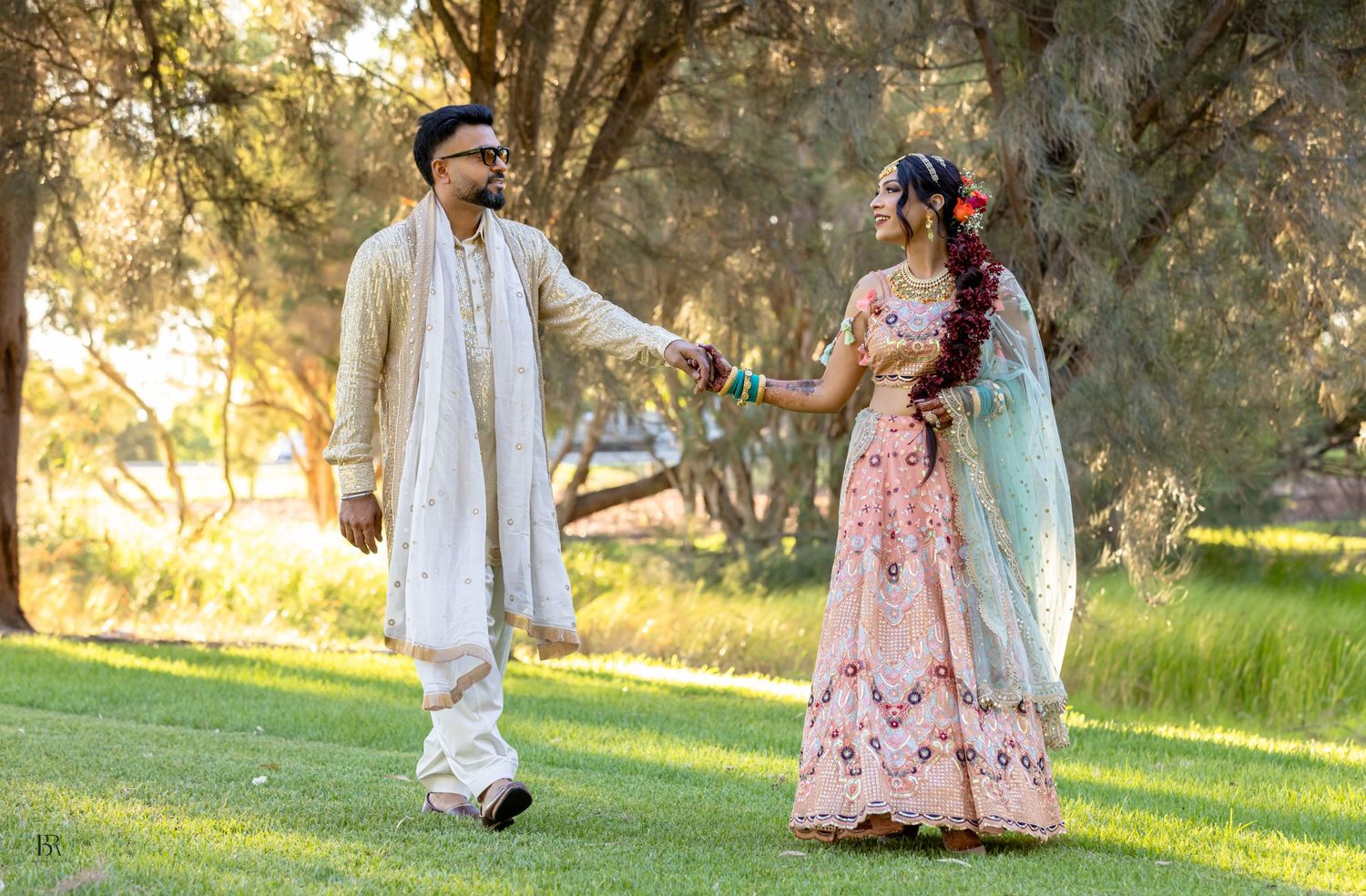 Indian wedding couple embracing in park, bride in pink lehenga and groom in traditional attire among eucalyptus trees.