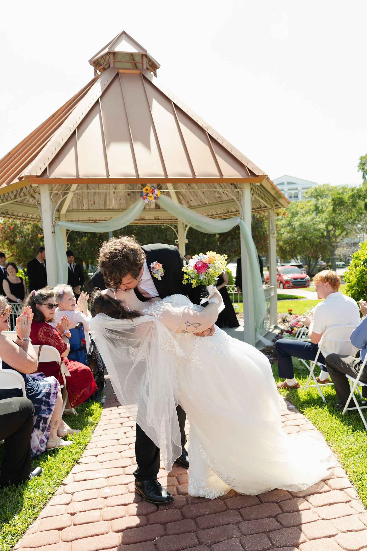 Groom dips bride in white gown for a romantic kiss on brick aisle outside gazebo at outdoor wedding ceremony.