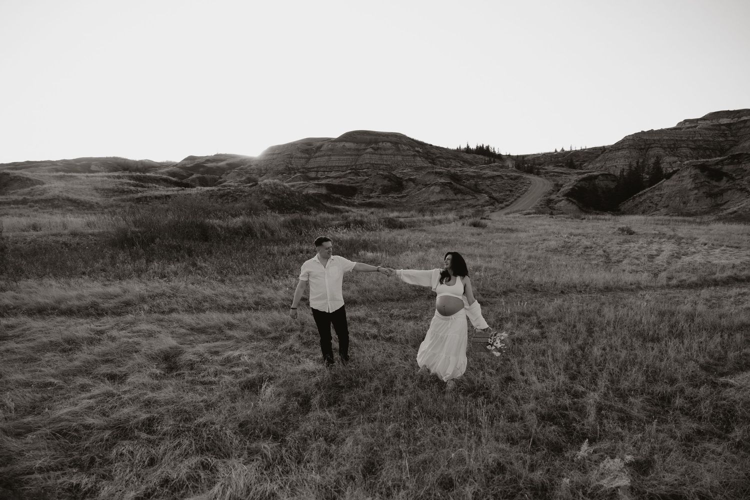 Black and white photo of a couple holding hands walking through a grassy field with rolling hills in the background.