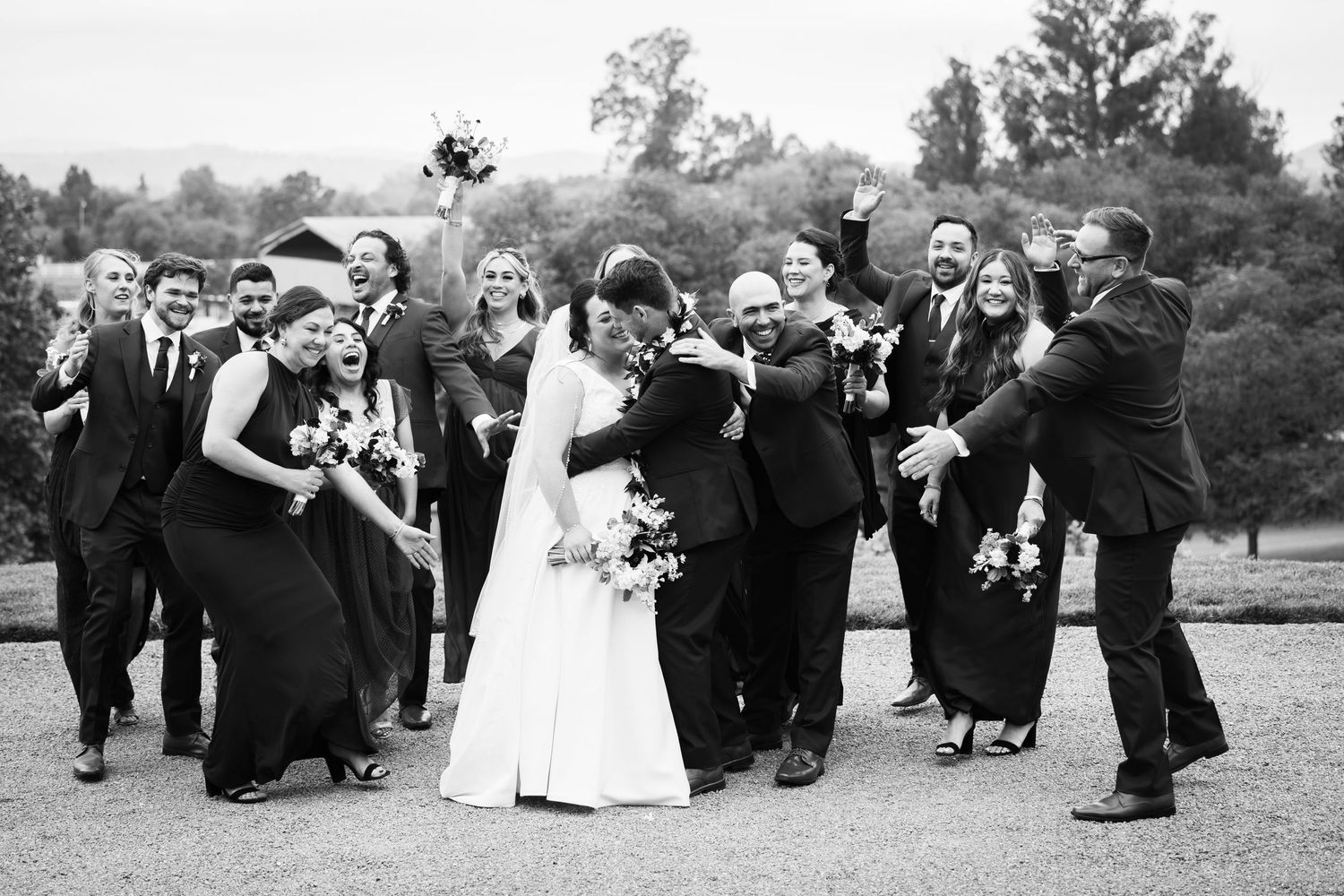 Black and white wedding party photo with bride and groom kissing surrounded by cheering guests outdoors.