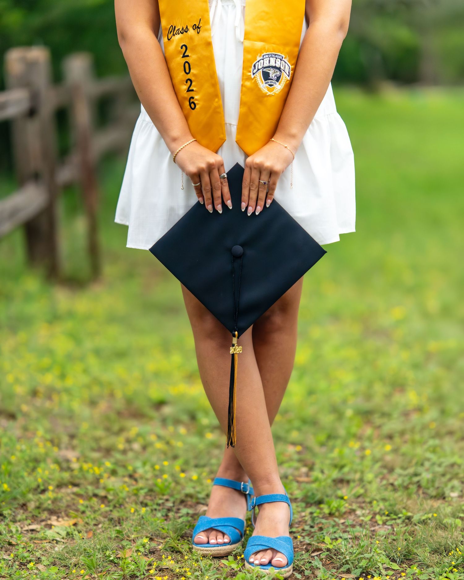 A Class of 2026 graduate holds a navy mortarboard cap wearing a gold stole and blue heels on a green lawn.