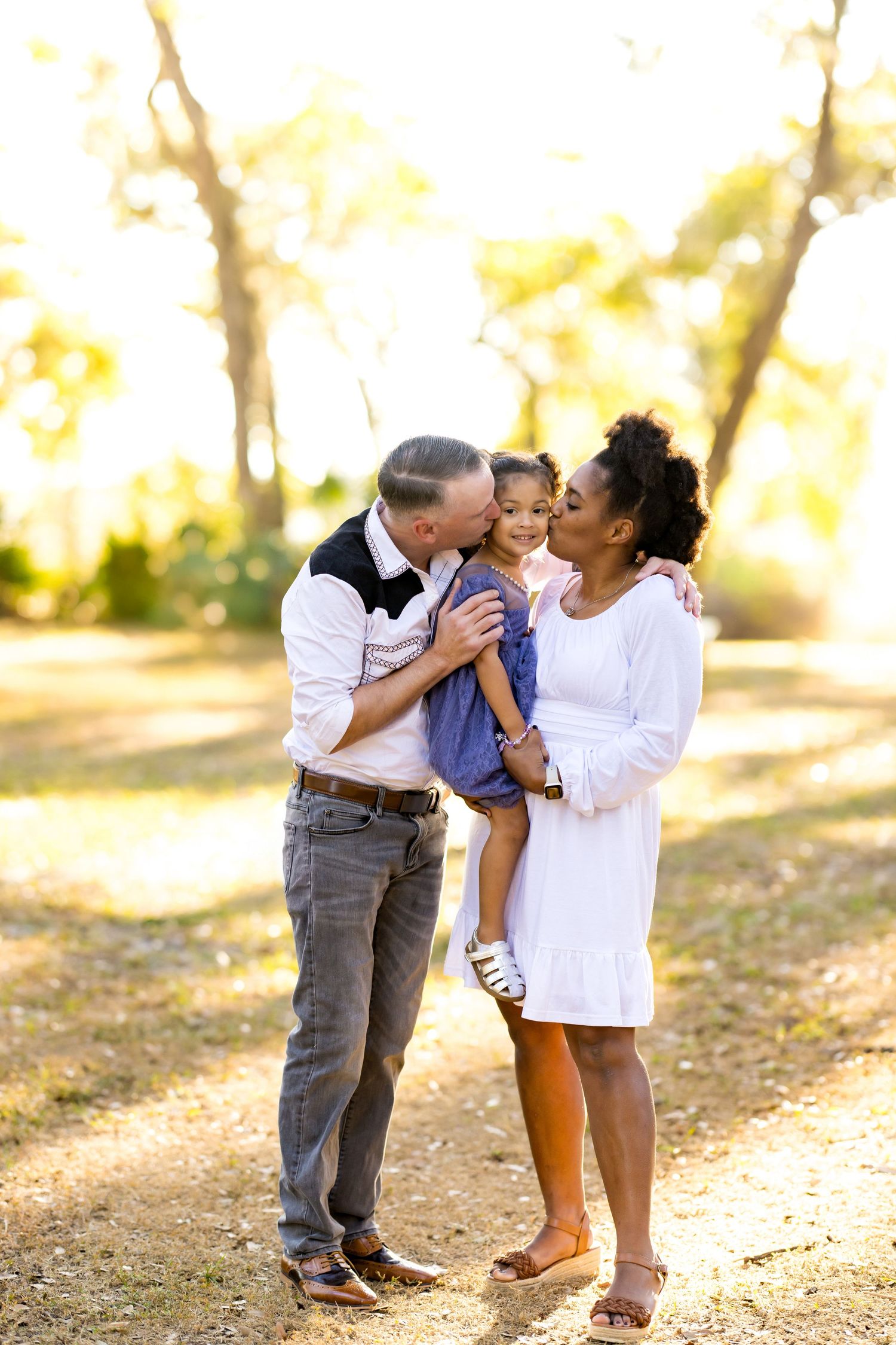A happy interracial family with a man and woman kissing their young daughter outdoors in a sunlit park.