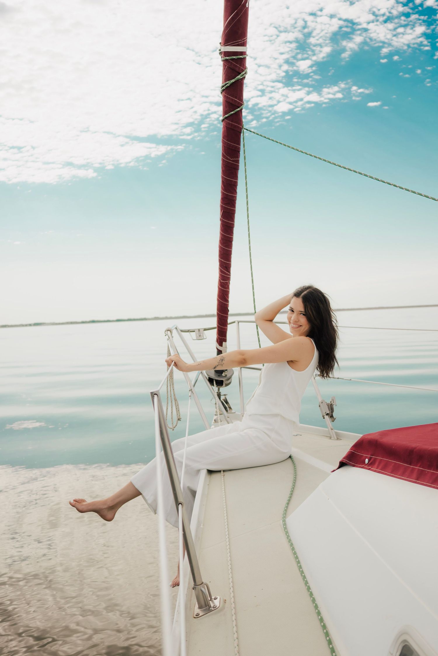 A smiling woman in white outfit relaxes on a sailboat deck, with calm blue water and cloudy sky in background.