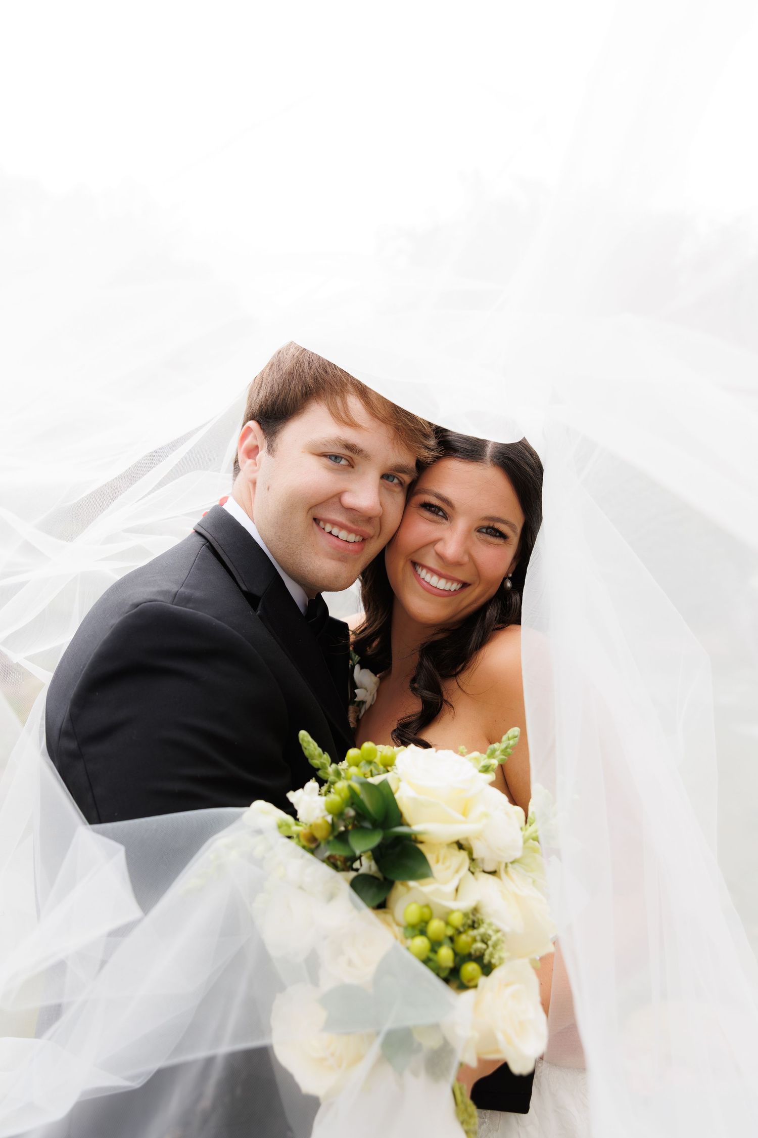 Newly married couple walking along brick path by white picket fence under tree canopy.