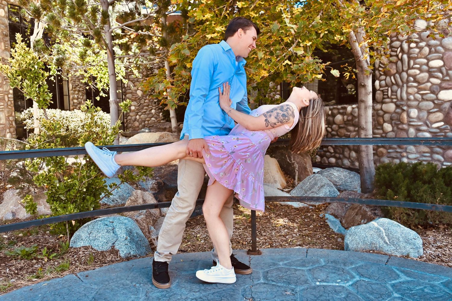 Couple dancing outdoors, man dipping woman in pink dress near stone wall with autumn trees.