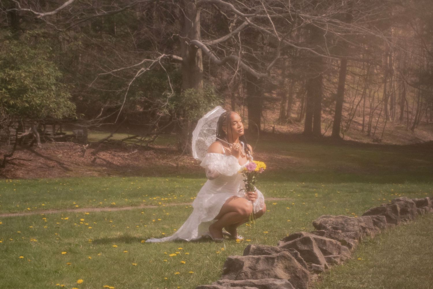 Bride in white wedding dress and veil kneeling on stone wall holding flowers in a misty forest setting.