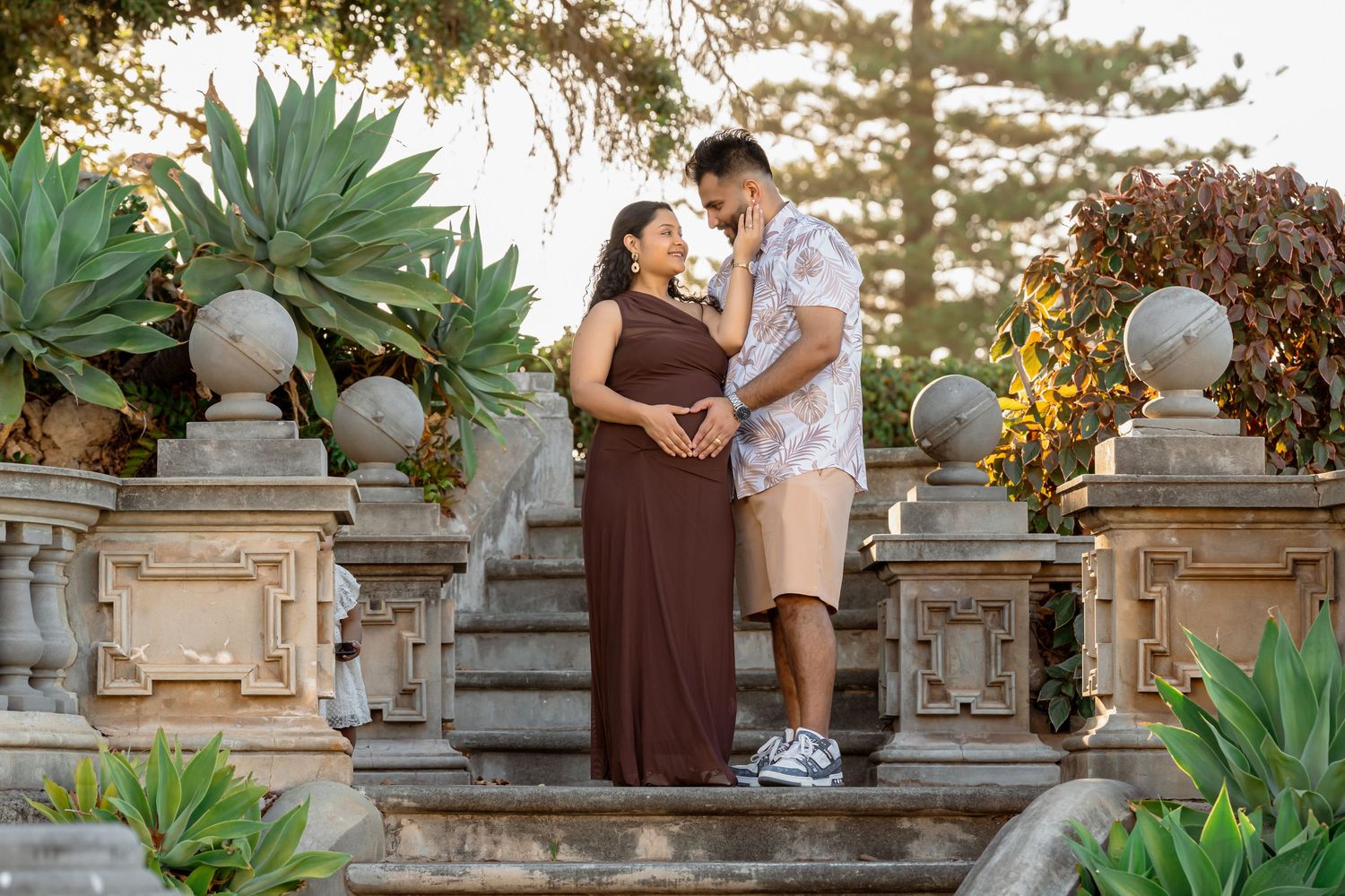 Couple sharing a tender maternity moment on stone steps surrounded by lush garden greenery in warm golden light.