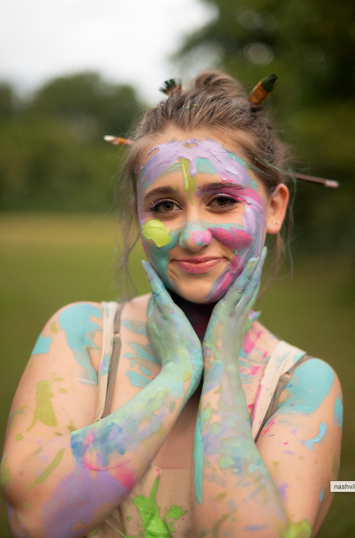 Young woman with colorful paint covering her face and hands, smiling outdoors with paintbrushes in her hair.