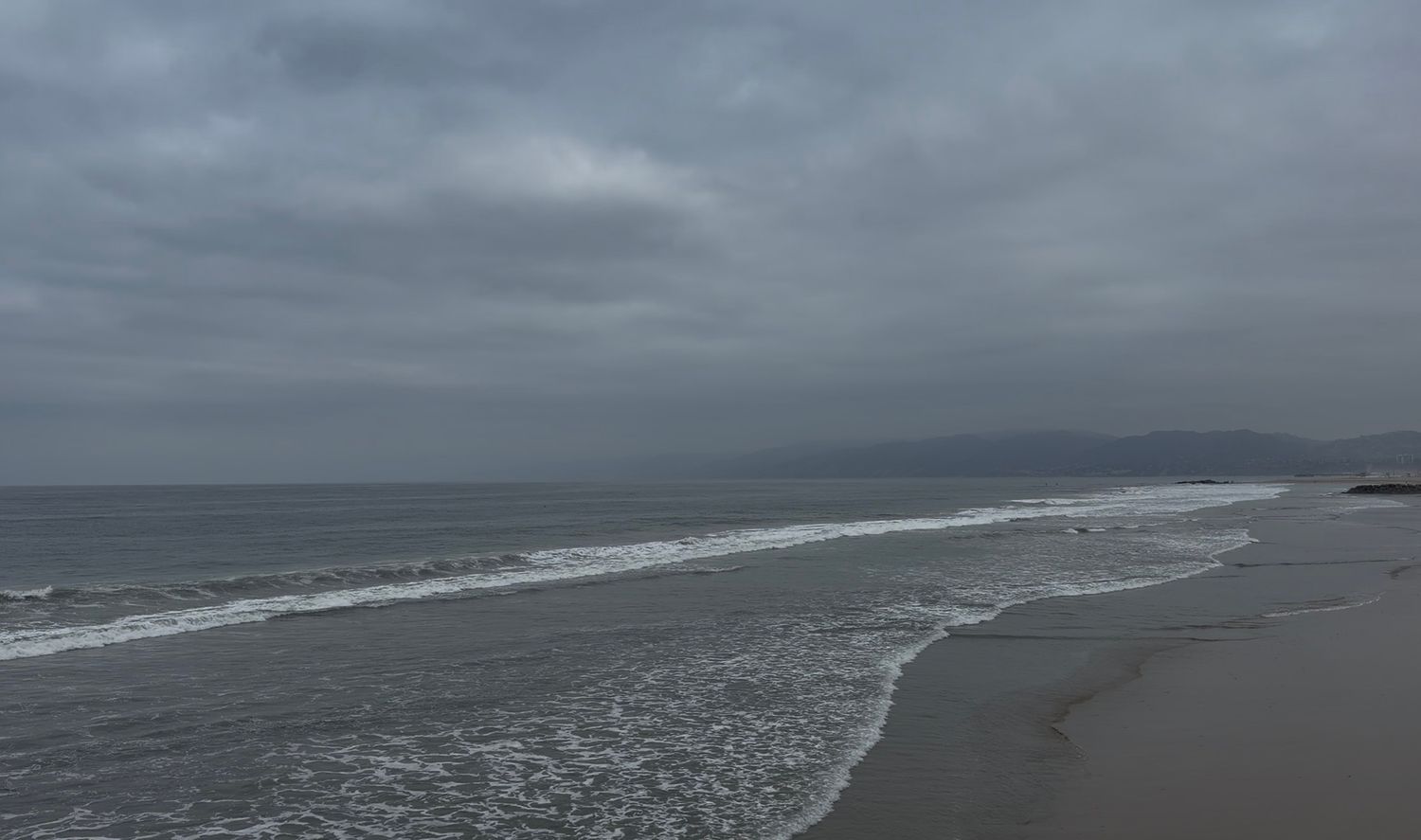 Overcast beach scene with gentle waves rolling onto a sandy shore under a cloudy gray sky with distant hills.