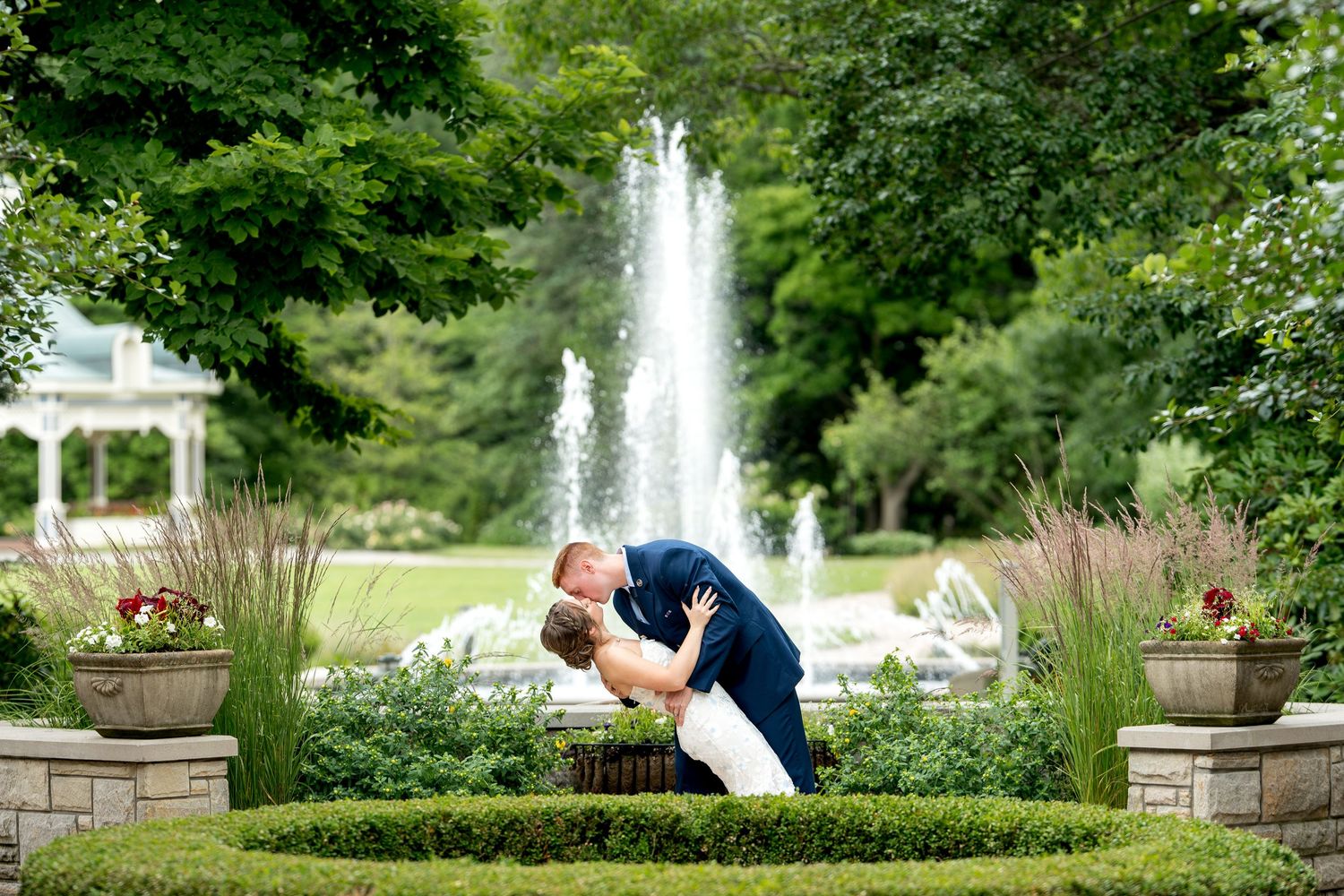 Couple sharing a romantic dip kiss in a lush garden with a fountain backdrop on their wedding day.