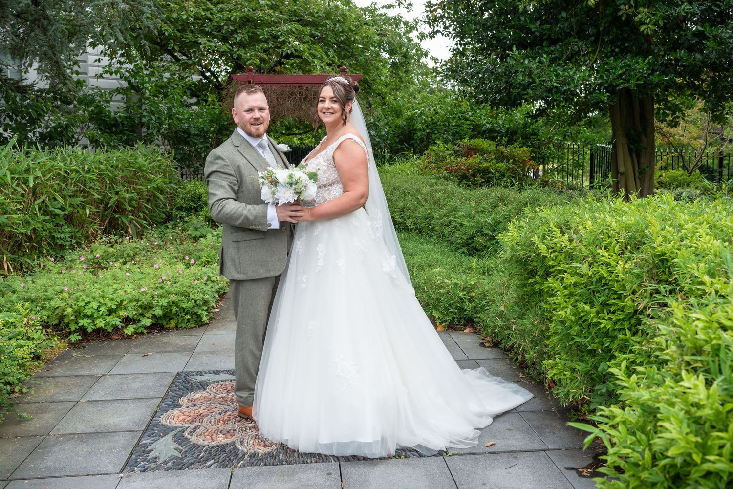 A bride and groom pose together in a lush garden, she in a white gown with veil, he in a grey suit.