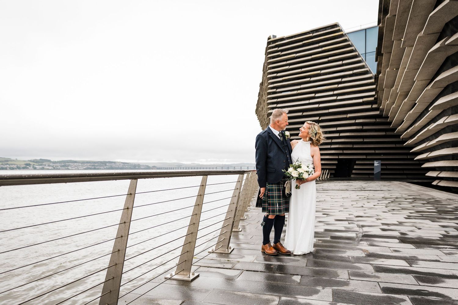 Wedding photo of bride in white and groom in kilt with V&A Dundee in background and Tay river.