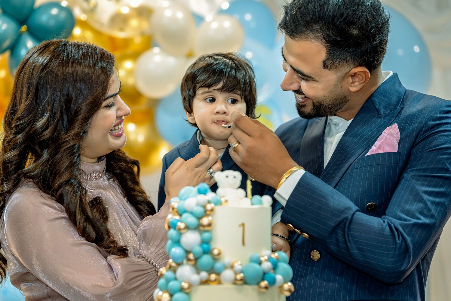 Family celebrates child's first birthday party with blue and gold balloon decorations and cake.